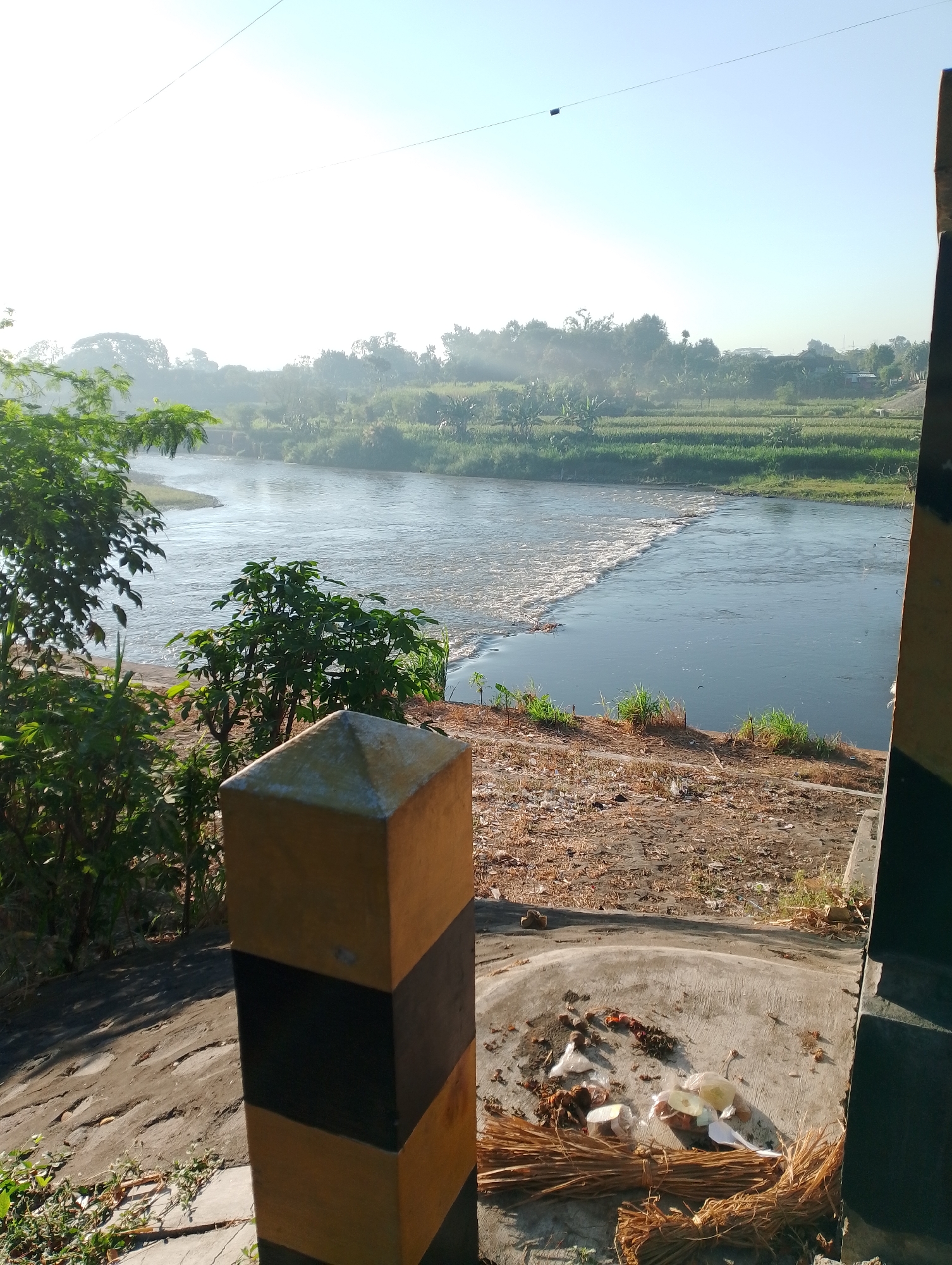 This is an outdoor, eye-level, vertical, medium shot of a river with agricultural fields in the background, taken on a clear, bright day. In the foreground, a black and yellow striped concrete post stands on the left side of the frame, casting a shadow. To the right of the post, a concrete area contains scattered debris, including what appears to be a broom made of dried grass and some food waste. A river flows from the left to the right, with a section of whitewater rapids in the middle, indicating a drop or obstruction. On the far bank of the river, lush green vegetation lines the water's edge, leading to a landscape of agricultural fields, possibly rice paddies, and scattered trees. In the distance, a hazy, light blue sky with a few faint power lines stretches across the horizon. The overall impression is a rural, possibly agricultural, setting in Mojo, Indonesia.