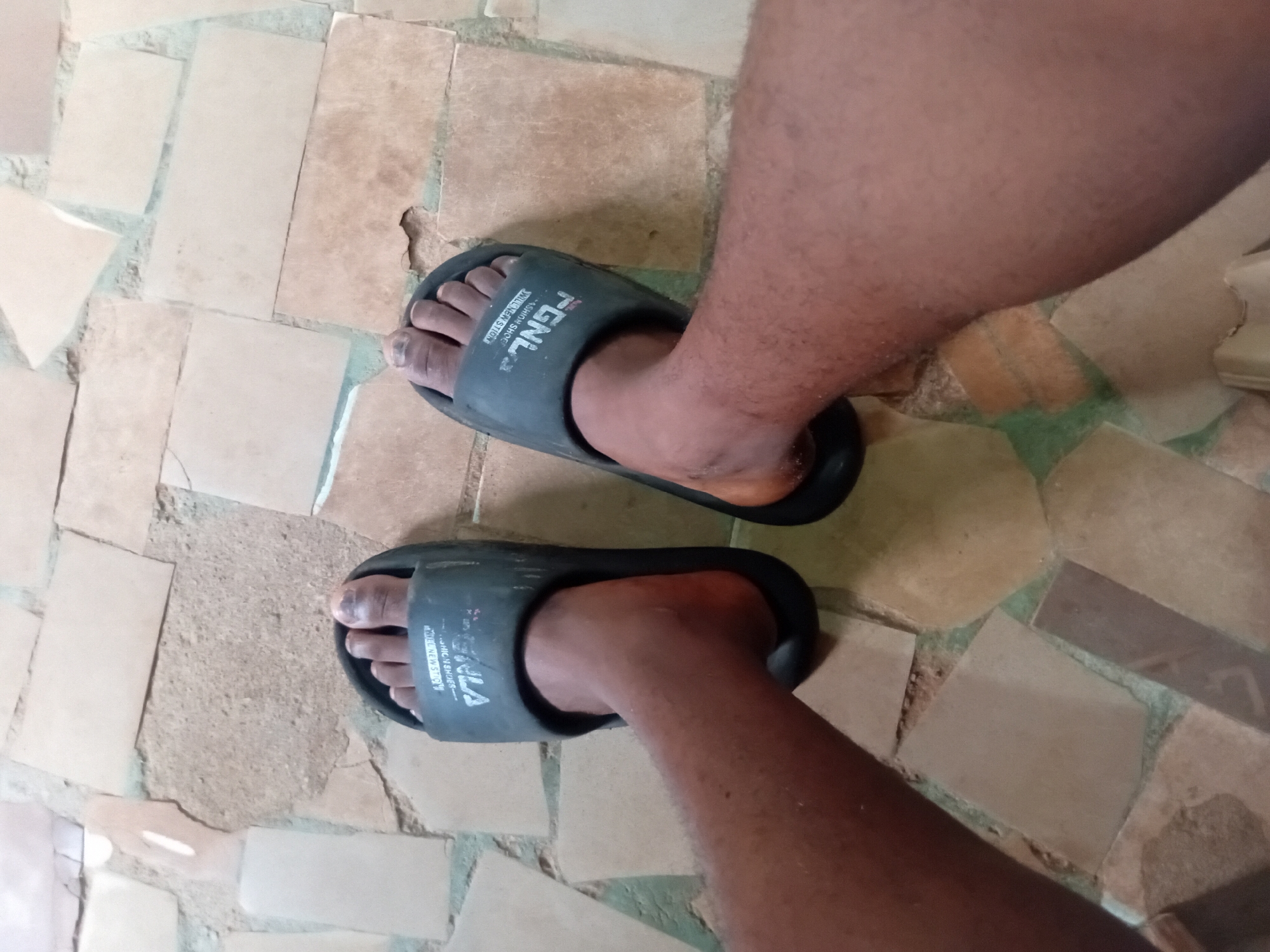 A pair of dark grey slide sandals, each with a white logo imprinted on the strap, are worn by an individual whose dark skin is visible on their feet and lower legs. The sandals are positioned on a floor composed of irregular, earth-toned tiles of varying sizes, interspersed with patches of grey grout. The scene appears to be indoors or in a covered outdoor area, given the tile flooring. There is no indication of the specific location within Nkpor, Nigeria, beyond the information provided. The image is taken from a top-down perspective, focusing on the feet and sandals.