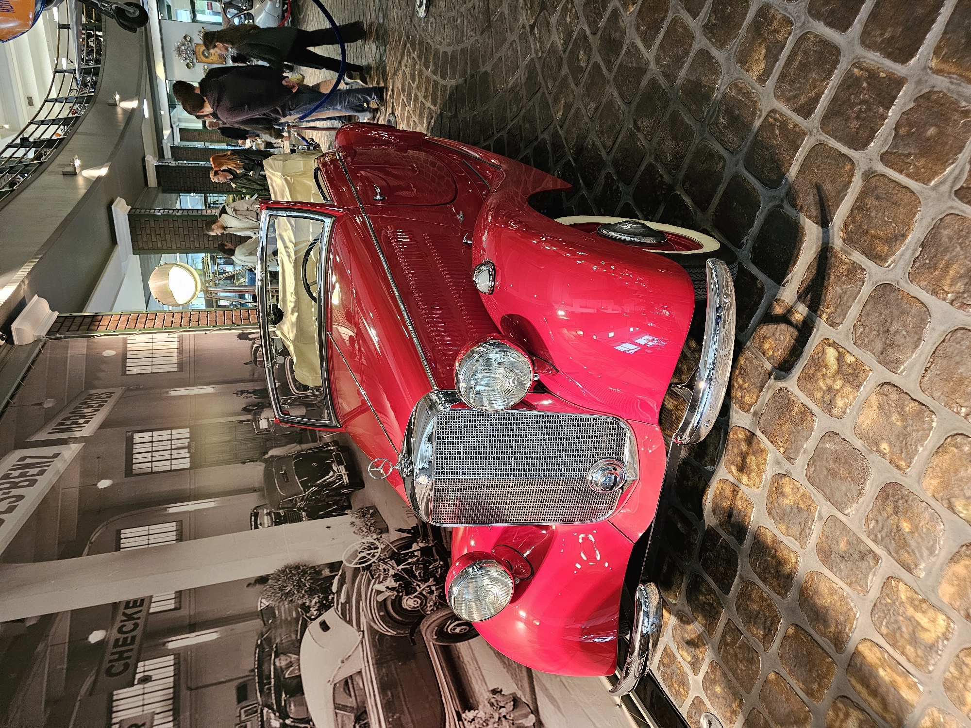 The image captures a vintage, bright red convertible car prominently displayed inside what appears to be a museum. The car is the main subject, with its chrome detailing and classic design capturing the eye. The background shows a mural featuring historical Mercedes-Benz vehicles and signage, indicating a car-related theme. To the left, there is a partial view of a balcony with a metal railing, suggesting an upper level of the museum. In the distance, indistinct figures of people are visible, likely museum visitors. The floor is paved with what appears to be cobblestone or a similar textured material. Given the information about Архангельское, Russia, it is plausible that this scene is inside a museum or exhibition within the Архангельское estate.