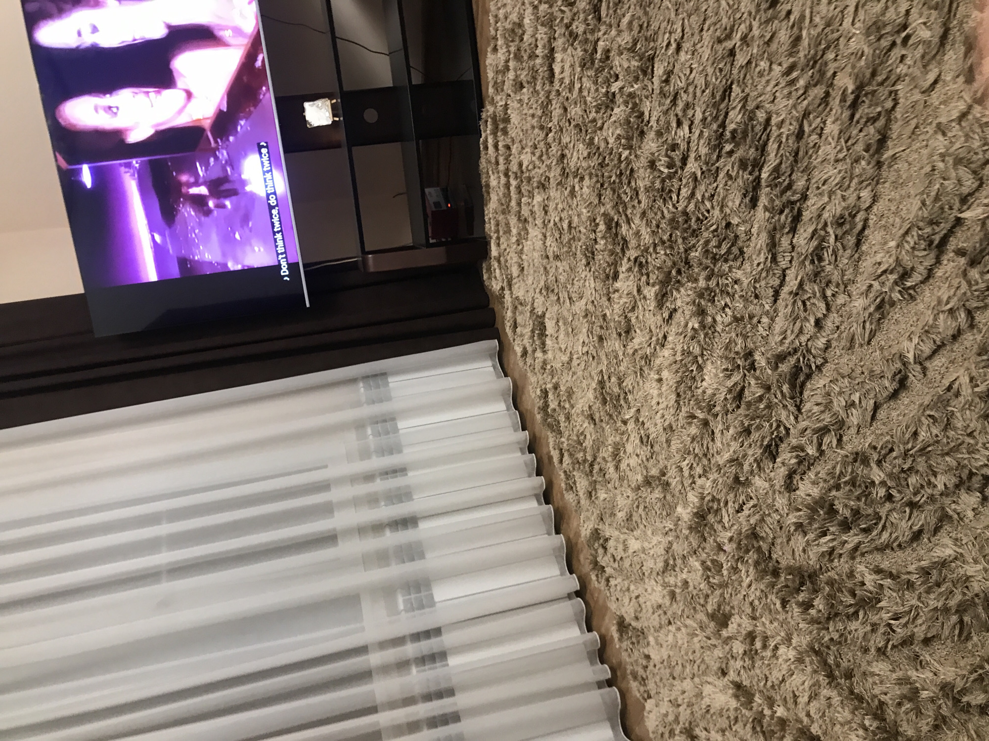 A dimly lit interior space featuring a television screen playing a video with captions, positioned next to a dark shelving unit. Beneath the television, window blinds hang. A thick, shaggy rug covers a large portion of the floor. There is no direct evidence to confirm the location is Yerevan, Armenia; the image lacks contextual clues connecting it to that city.