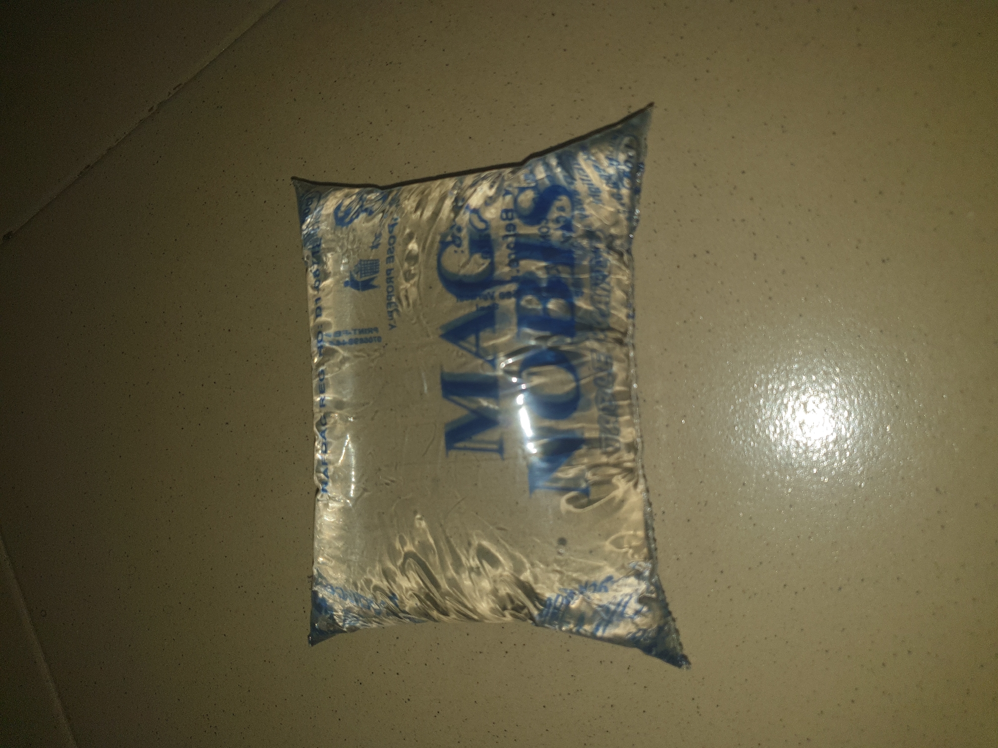 A sealed sachet of water, branded "MAG NOBIS", is resting on a speckled tiled surface. The sachet is transparent with blue text and graphics. The surface appears to be a floor or countertop. The lighting suggests an indoor environment, possibly at night or in a dimly lit room. The overall scene is static and unremarkable, with no visible people or activity. Based on the provided context, this scene is located in Jalingo, Nigeria.