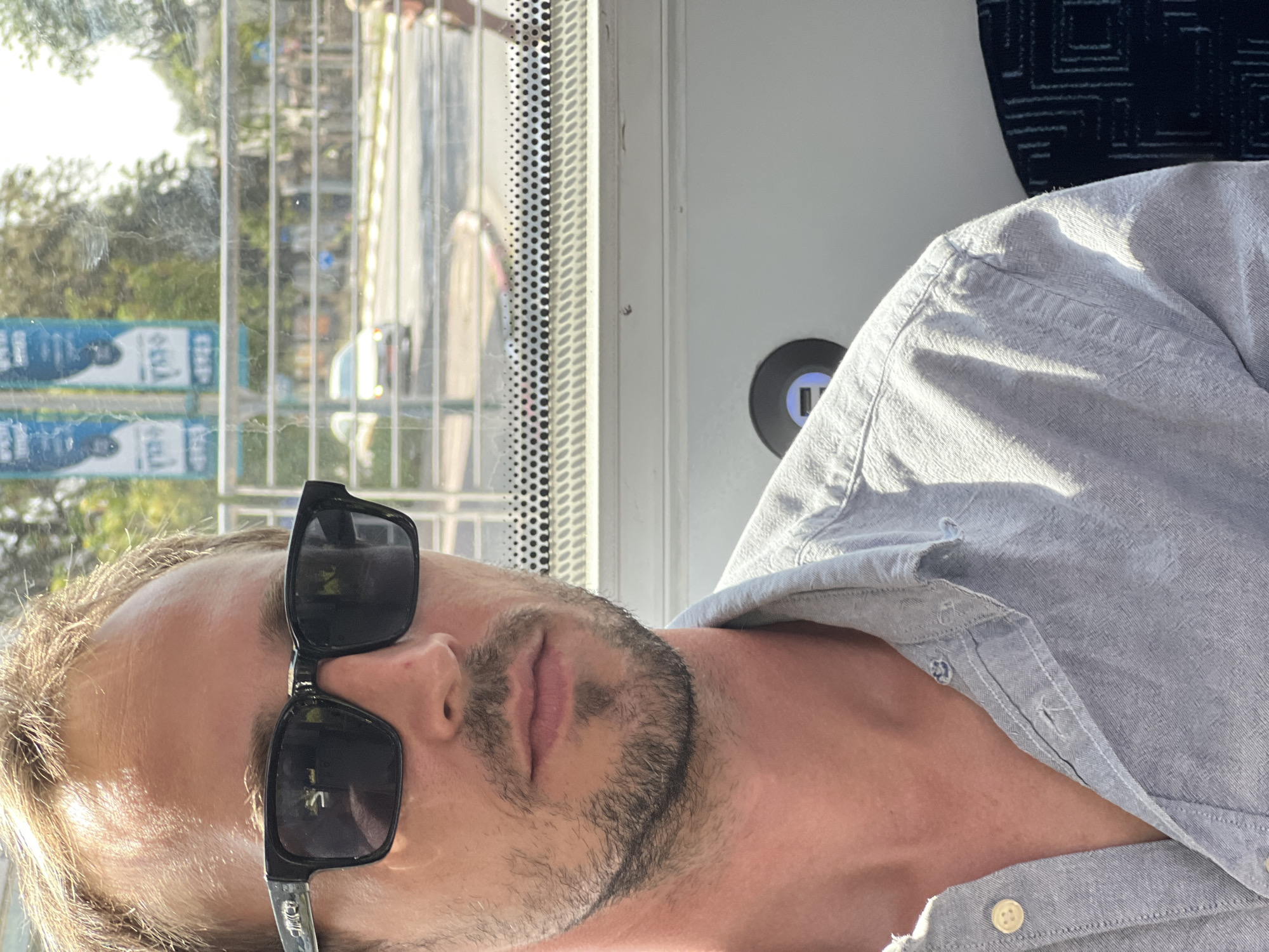 A close-up, selfie-style photo shows a man with light brown hair, wearing dark sunglasses and a light blue button-down shirt. He has stubble on his face. The man is seated indoors, possibly on public transportation, as indicated by the window with horizontal blinds and a view of a street scene outside. The exterior view includes cars, trees, and what appear to be street banners with text in Hebrew, suggesting the location is Kiryat Bialik, Israel. Inside the vehicle, a black circular object with what looks like USB ports is visible on the wall. The man's left shoulder and arm are visible in the lower left corner of the frame, and a patterned seat is partially visible on the right. The lighting suggests it is daytime and sunny.