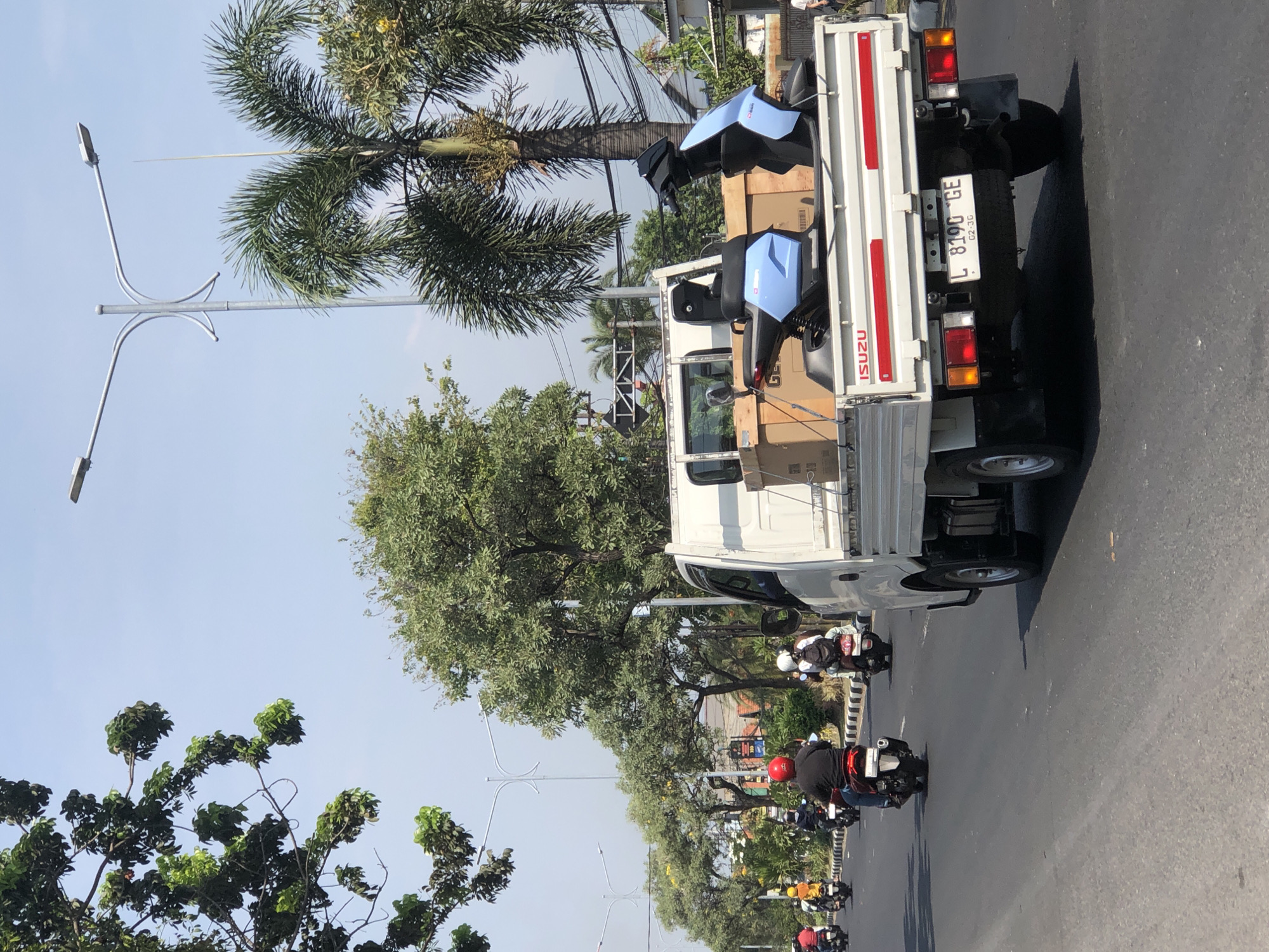 In the city of Larangan, Indonesia, a white Isuzu flatbed truck is transporting two light blue electric scooters on its bed. The truck is traveling on a paved road. Several motorcyclists are also on the road, heading in the same direction as the truck. The environment appears to be urban or suburban, with trees and palm trees lining the roadside. Overhead, streetlights are visible. The sky is clear and blue, indicating a sunny day. The truck has a license plate that reads "L 8190 GE". One of the electric scooters is positioned at the front of the truck's bed, while the other is at the back. Cardboard boxes are also visible on the truck bed, near the scooters. The scene is captured from a slightly elevated perspective, looking down and to the right.