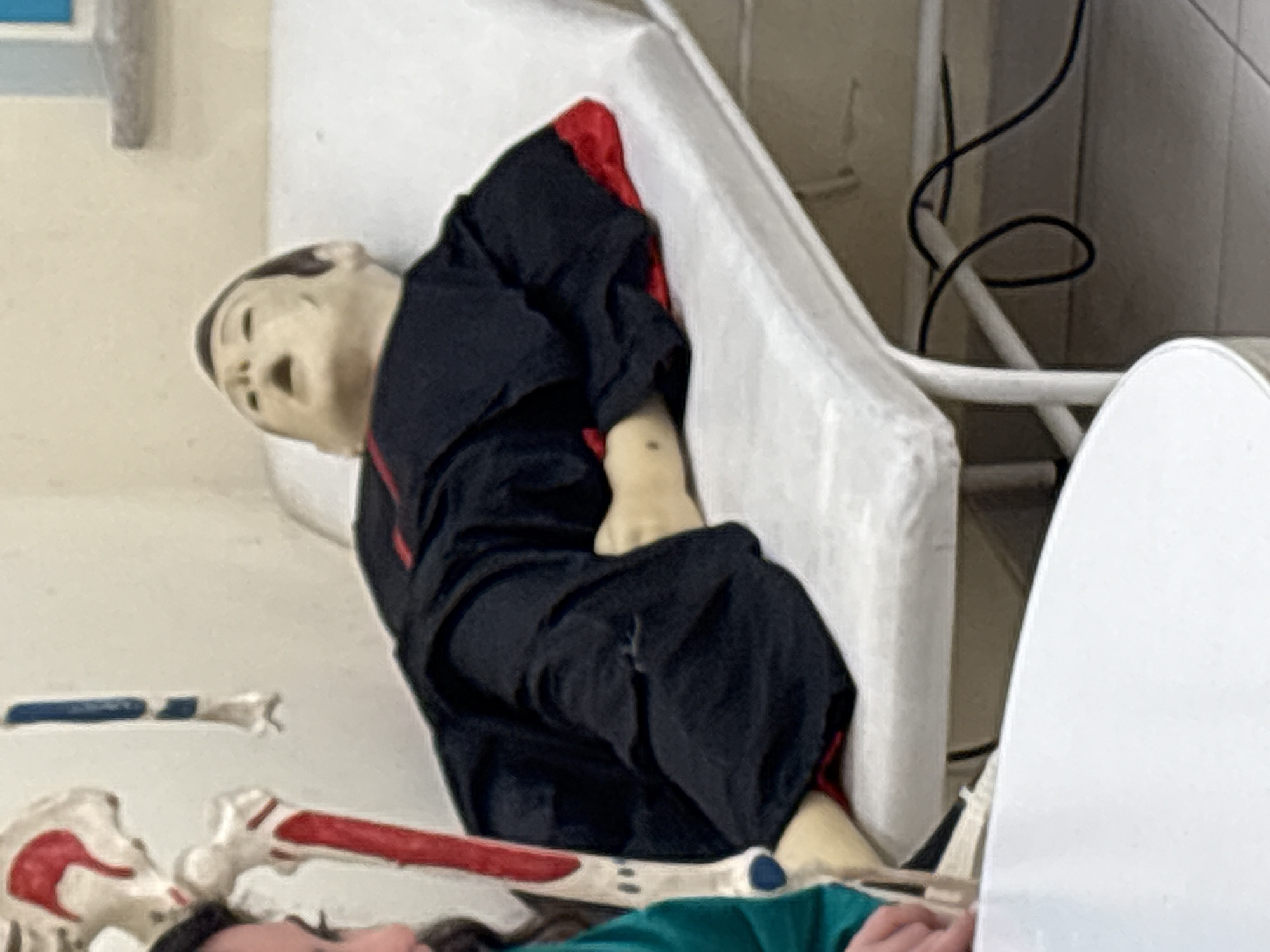 The image depicts a medical training setting, likely a classroom or laboratory in Yerevan, Armenia. In the foreground, a white table occupies the lower left portion of the frame. A mannequin dressed in black clothing with red trim is lying on a white examination table. The mannequin is positioned on its back with its head tilted back and mouth open. To the left of the mannequin, a partial view of a human skeleton is visible, with red and blue accents on its bones. The background is a plain, light-colored wall. Beneath the examination table, a black electrical cord is visible, suggesting the presence of medical equipment. A person's hand, partially visible on the far left, is also present, suggesting the scene is being observed or interacted with.