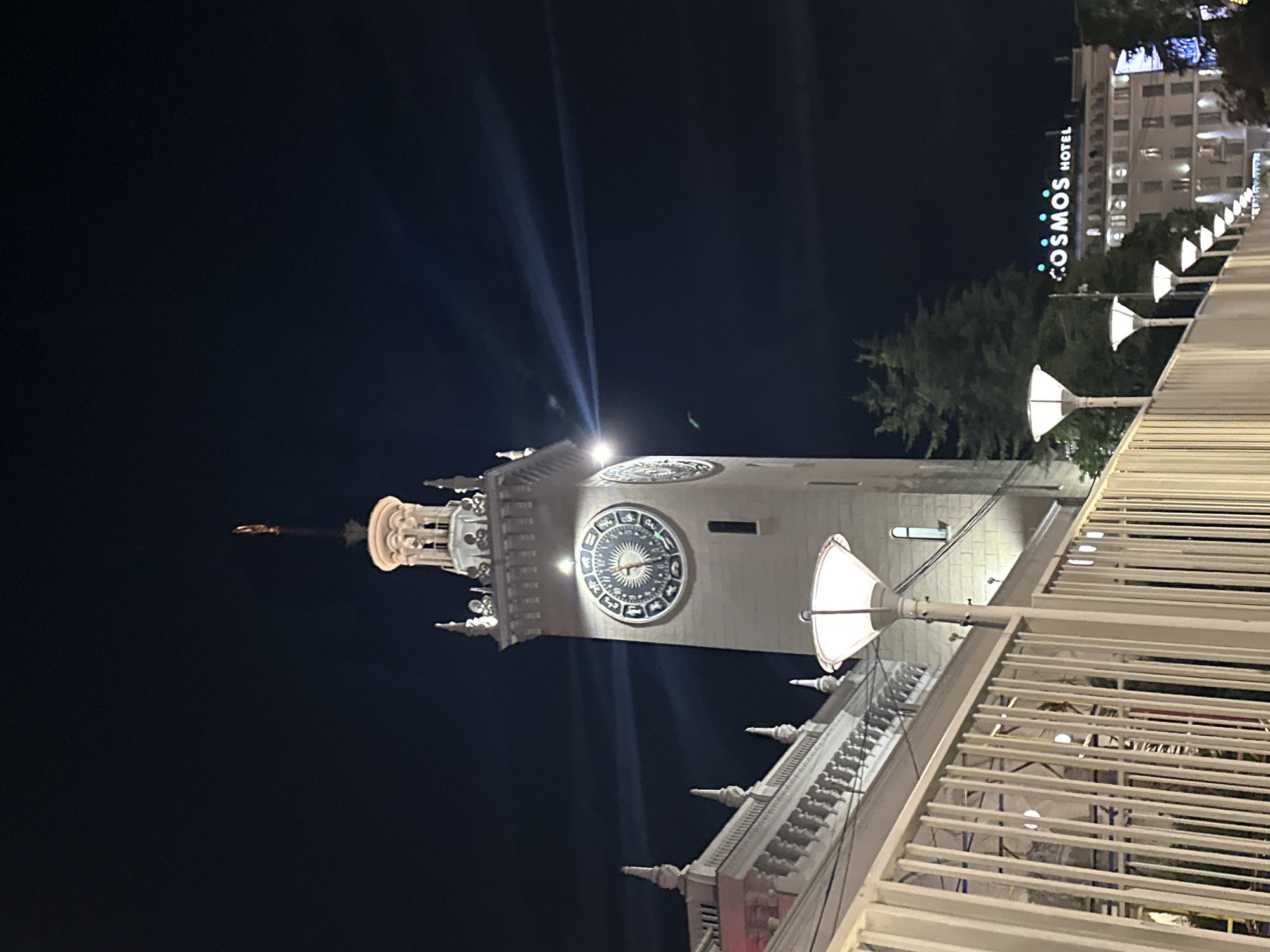 The image shows a night view of the Sochi Railway Station in Sochi, Russia. The station's clock tower, illuminated and featuring a zodiac dial, is prominently visible. In the foreground, a white metal fence lines a pathway with modern lampposts casting a glow. Behind the fence, a white ornate structure, likely part of the station building, is visible. Further in the background, a building with the illuminated sign "COSMOS HOTEL" is present. Several evergreen trees are interspersed between the buildings. The sky is dark, with some faint light beams emanating from the clock tower.