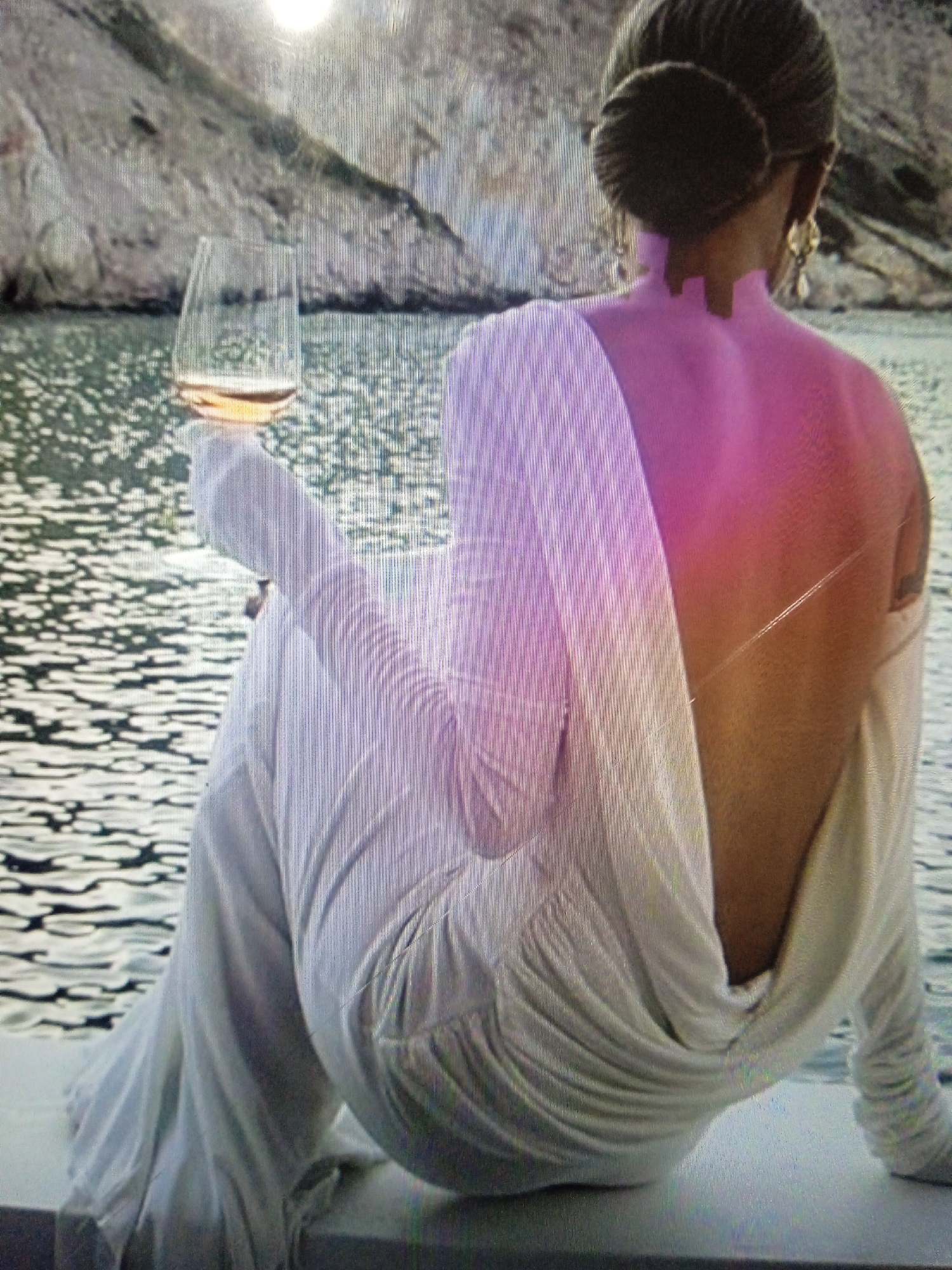 The image shows a person from behind, wearing a white draped dress and sitting near a body of water. A glass of rose wine is held by the person. The background features a rocky cliff and a bright sun. The person's back is exposed and highlighted in a pink hue. This image does not contain any elements or context that would place it in Chernivtsi, Ukraine.