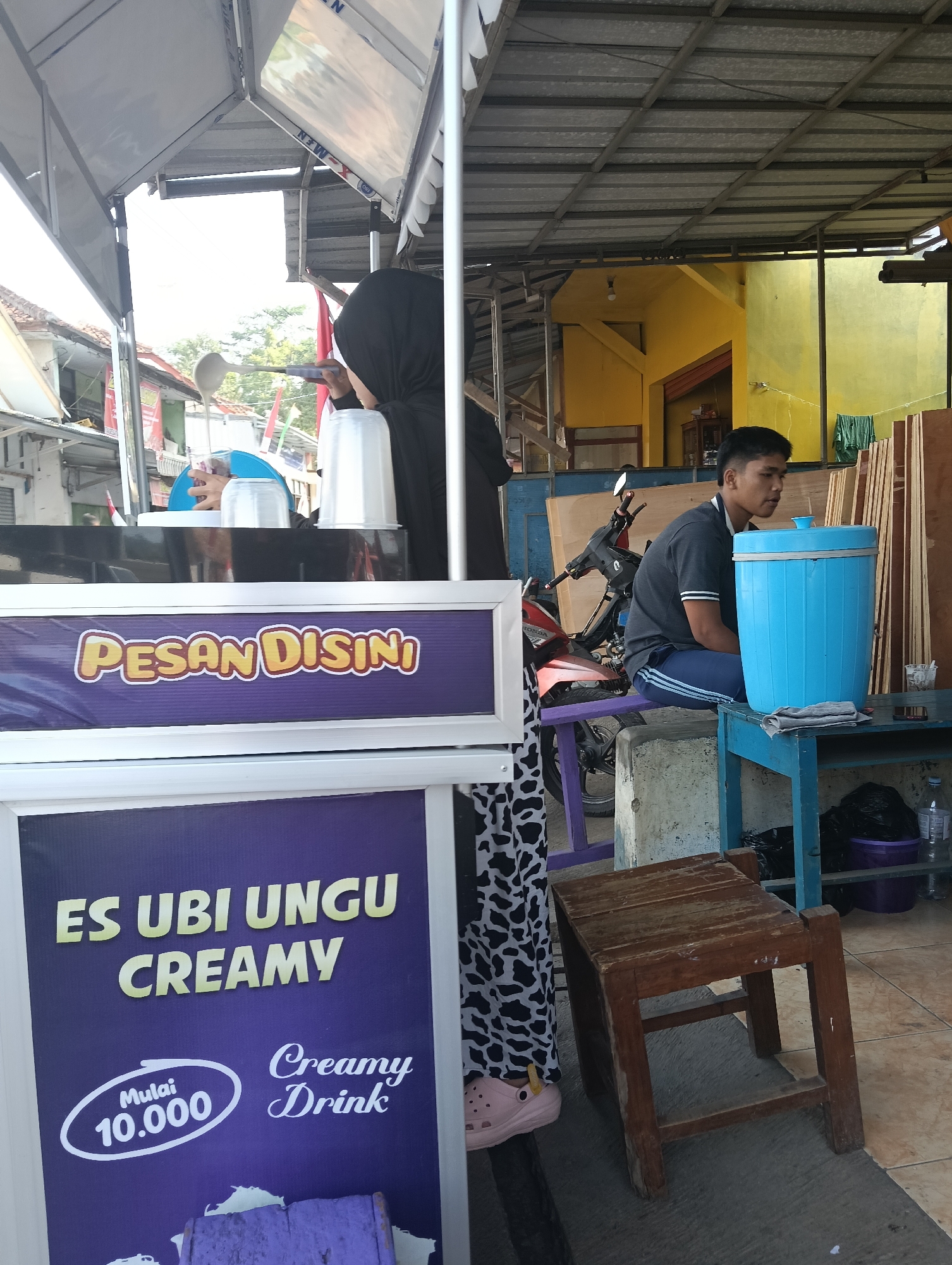 The image depicts an outdoor scene, likely a roadside stall or small shop, with a prominent purple sign that reads "PESAN DISINI" (Order Here) in yellow, stylized lettering. Below this, another purple sign displays "ES UBI UNGU CREAMY" (Purple Sweet Potato Creamy Ice) in large yellow letters, with additional text indicating "Mulai 10.000" (Starting from 10,000) and "Creamy Drink" in white.
In the foreground, a person wearing a black hijab and a patterned black and white garment is partially visible, holding a ladle over a white container. To the right, a young man wearing a dark grey t-shirt and blue track pants is seated on a blue bench, looking to his right. Beside him is a large, bright blue cylindrical container. A small, rustic wooden stool is positioned in front of the bench. The background reveals a yellow building and other structures, suggesting an urban or semi-urban environment. A red and black motorcycle is parked behind the bench where the young man is seated. The ground appears to be tiled or paved. The overall impression is of a food or beverage stall in Puspahiang, Indonesia, potentially focusing on a local specialty.