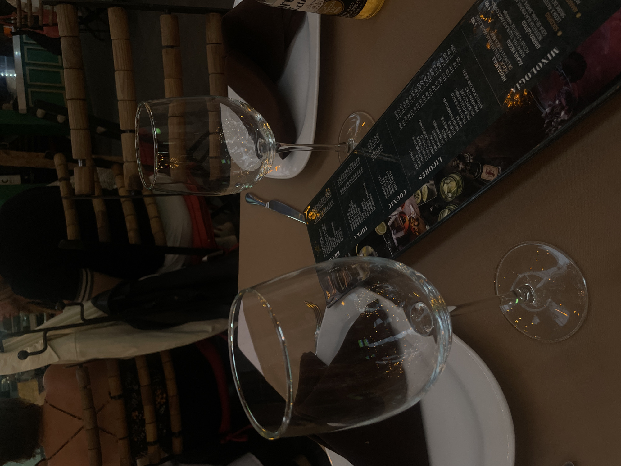 The image shows a table setting at a restaurant in Puebla, Mexico, with two empty wine glasses and a partially visible menu. A beer bottle is also present, along with plates and cutlery. A person is seated at the table. The environment suggests an indoor dining setting.
