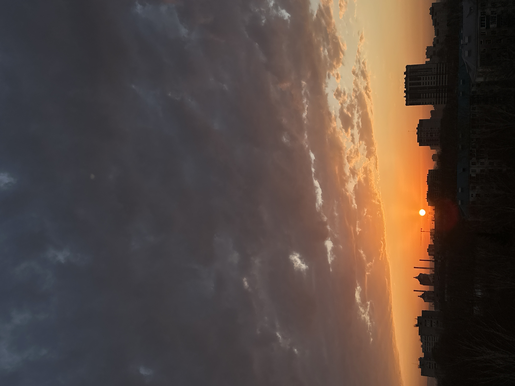 The image captures a vivid sunset over the city of Kazan, Russia. In the foreground, a dark silhouette of buildings and trees forms the lower edge of the frame. The setting sun casts a warm orange glow that dominates the sky. The color transitions into a lighter, almost yellow hue near the horizon. Clouds are present on both sides of the sky, ranging from dark, ominous tones to lighter, sunlit shades. A construction crane is visible near the horizon. The city skyline is punctuated by tall buildings.