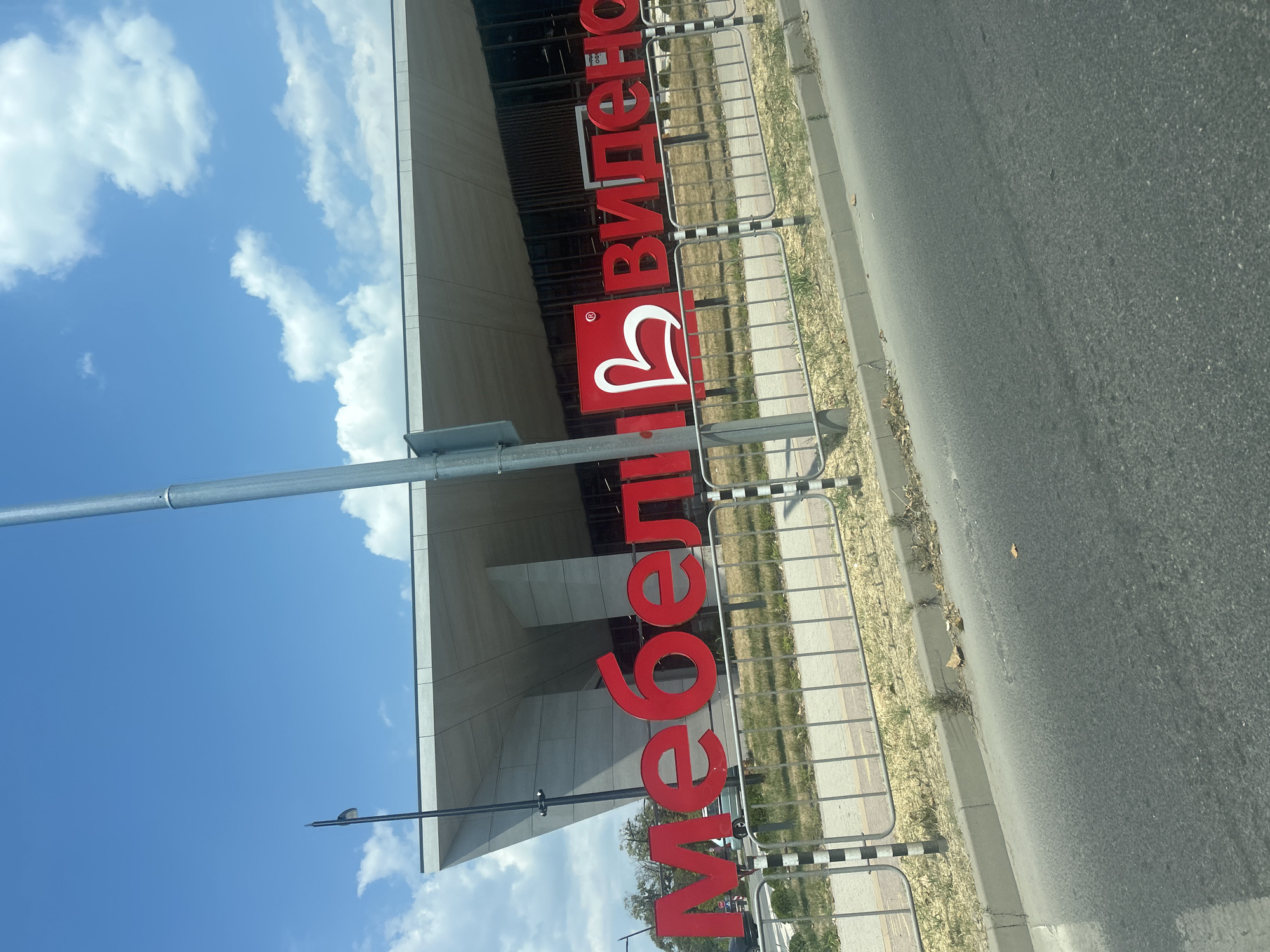 The image shows the exterior of a modern building with a large red sign that reads "МЕБЕЛІ ВІДЄНО". The building has a large, cantilevered roof with a gray facade. In front of the building is a metal fence with black and white striped posts. The sign is made up of large, red, capital letters, with a white heart shape integrated into the design of the second word. The background features a bright blue sky with scattered white clouds. The scene is observed from a roadside perspective, with a portion of an asphalt road and a sidewalk visible in the foreground. The location is identified as the Möbel Vidjeno store in Plovdiv, Bulgaria.