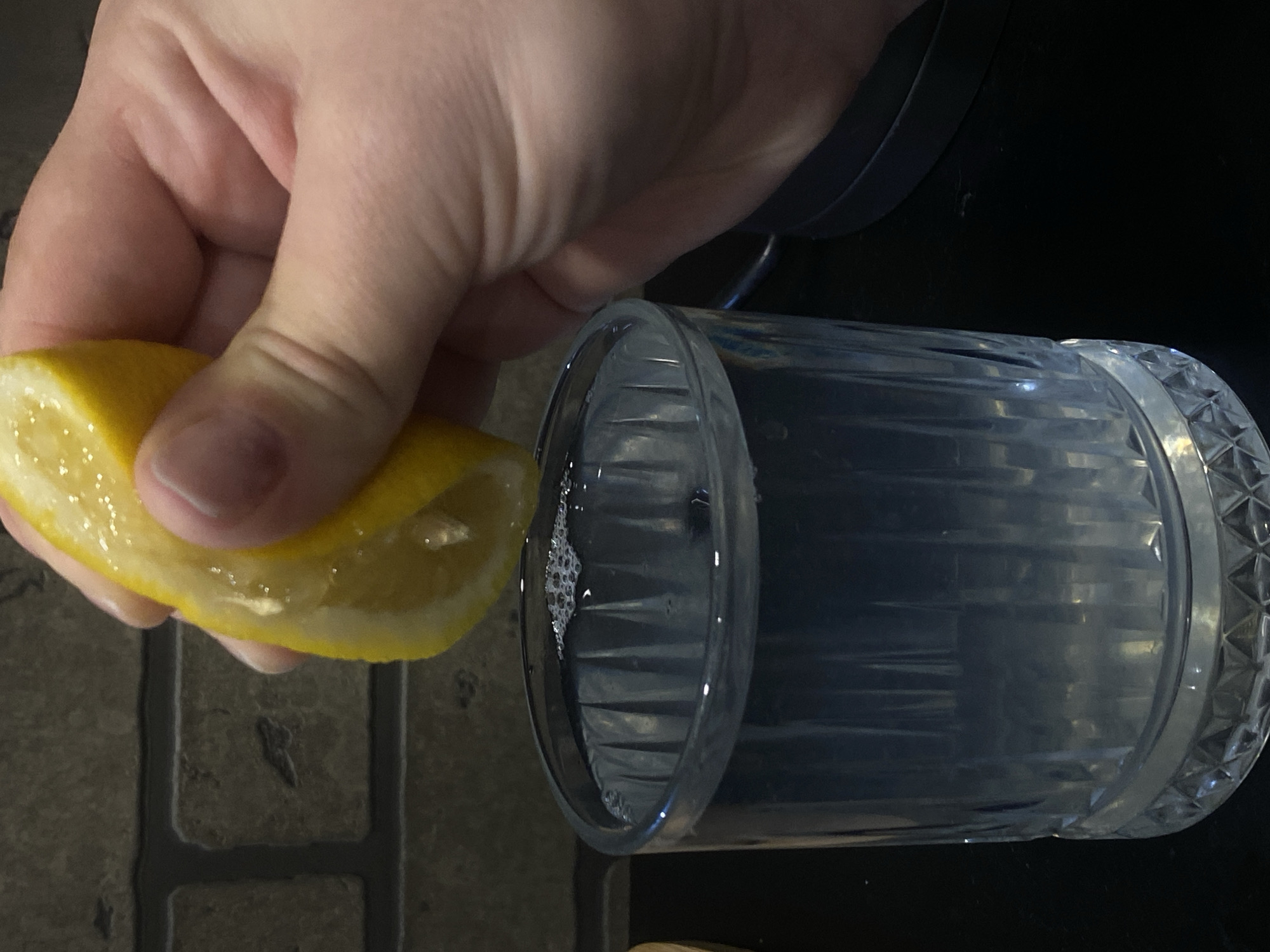 A person's hand is squeezing a lemon wedge into a crystal glass filled with a clear liquid. The background is dark with a textured surface visible underneath. The overall scene appears to be indoors. The location is not apparent from the image and cannot be determined to be Sochi, Russia.