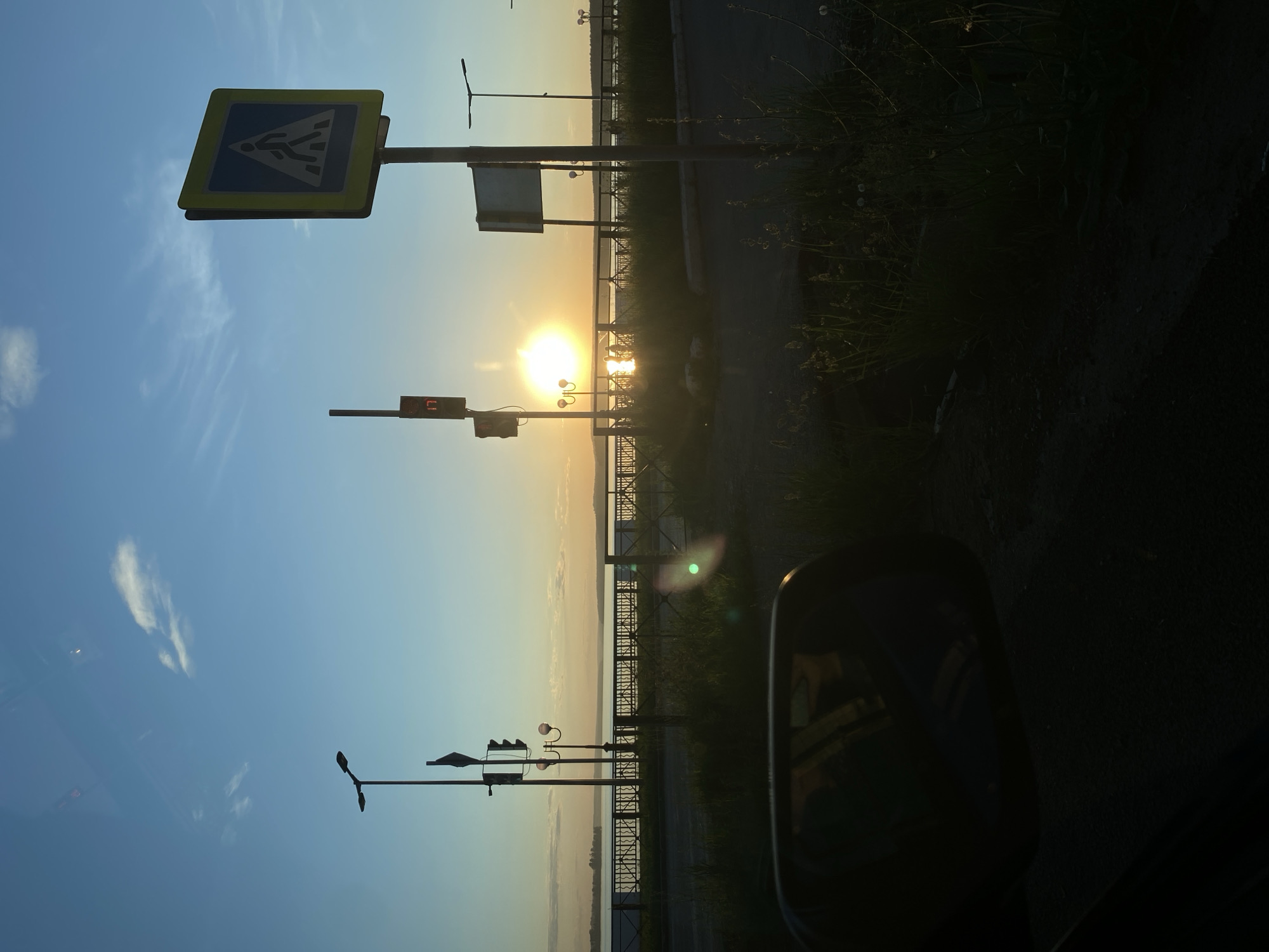 The image presents a backlit view of a waterfront area at sunrise or sunset, situated in Votkinsk, Russia. The perspective is from within a vehicle, evidenced by a dark car side mirror visible in the lower-left quadrant, reflecting faint internal details.
In the foreground, a dark surface of asphalt and overgrown dry grass covers the lower section of the frame. A decorative black metal fence, featuring a recurring crisscross pattern, extends horizontally across the mid-ground, acting as a barrier between the foreground and the water's edge.
Behind the fence, various urban infrastructure elements are silhouetted. On the left, a tall street lamp post with two luminaires stands alongside a pole supporting a traffic light and a red digital countdown display showing '17'. To the right, a square road sign with a yellow border and a blue interior depicts a white pedestrian crossing symbol. Further right, a blank rectangular sign and another single-luminaire street lamp are visible. Several ornate street lamps with spherical tops line the embankment along the water. Multiple indistinct human figures are discernible in silhouette on the promenade behind the fence, some interacting with the bright sun reflection on the water.
The background is dominated by a large, calm body of water, identified as the Votkinsk Reservoir, reflecting the intense light of the low-lying sun. The sun itself appears as a brilliant, circular disc with visible lens flare. A distant, dark landmass, likely the opposite bank of the reservoir, stretches across the horizon. The sky transitions from a deep blue at the zenith to a warm golden hue near the horizon, populated by a few scattered white clouds. Faint reflections, possibly of clouds or vehicle interior, are discernible on the upper portion of the image.