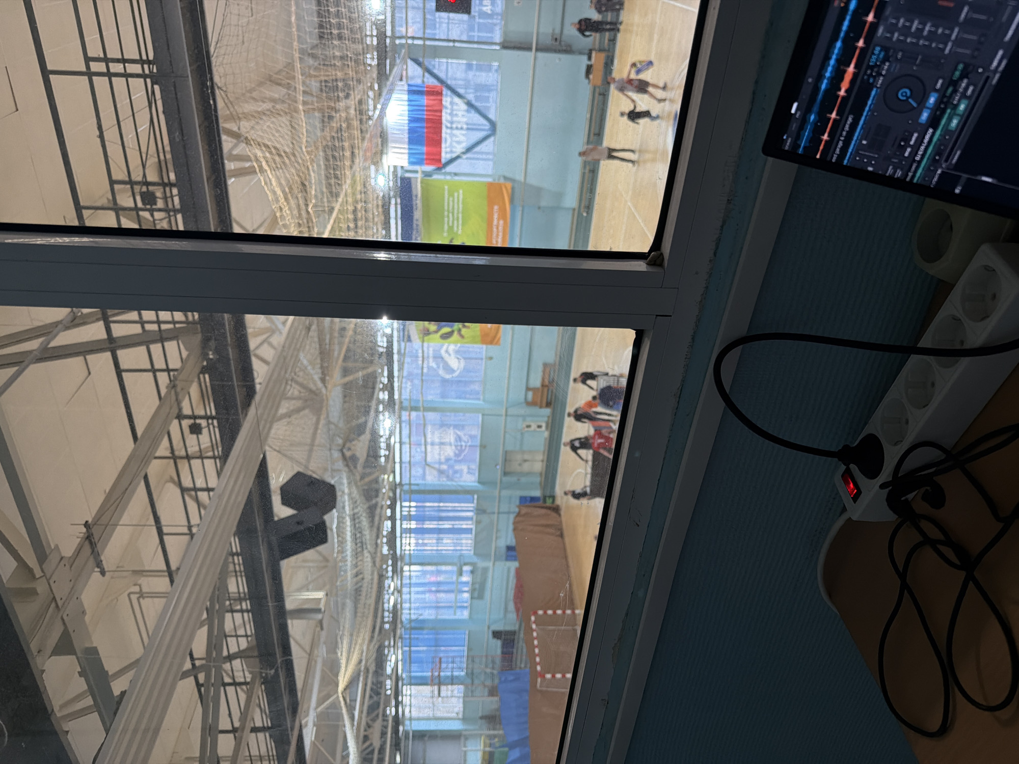 A view from a window overlooking an indoor sports arena in Кондратово, Russia. Inside the arena, people are moving around on the court, and the Russian flag is visible. A laptop showing DJ software, an electrical outlet strip, and power cords are visible in the foreground. The arena has high ceilings with metal support beams.