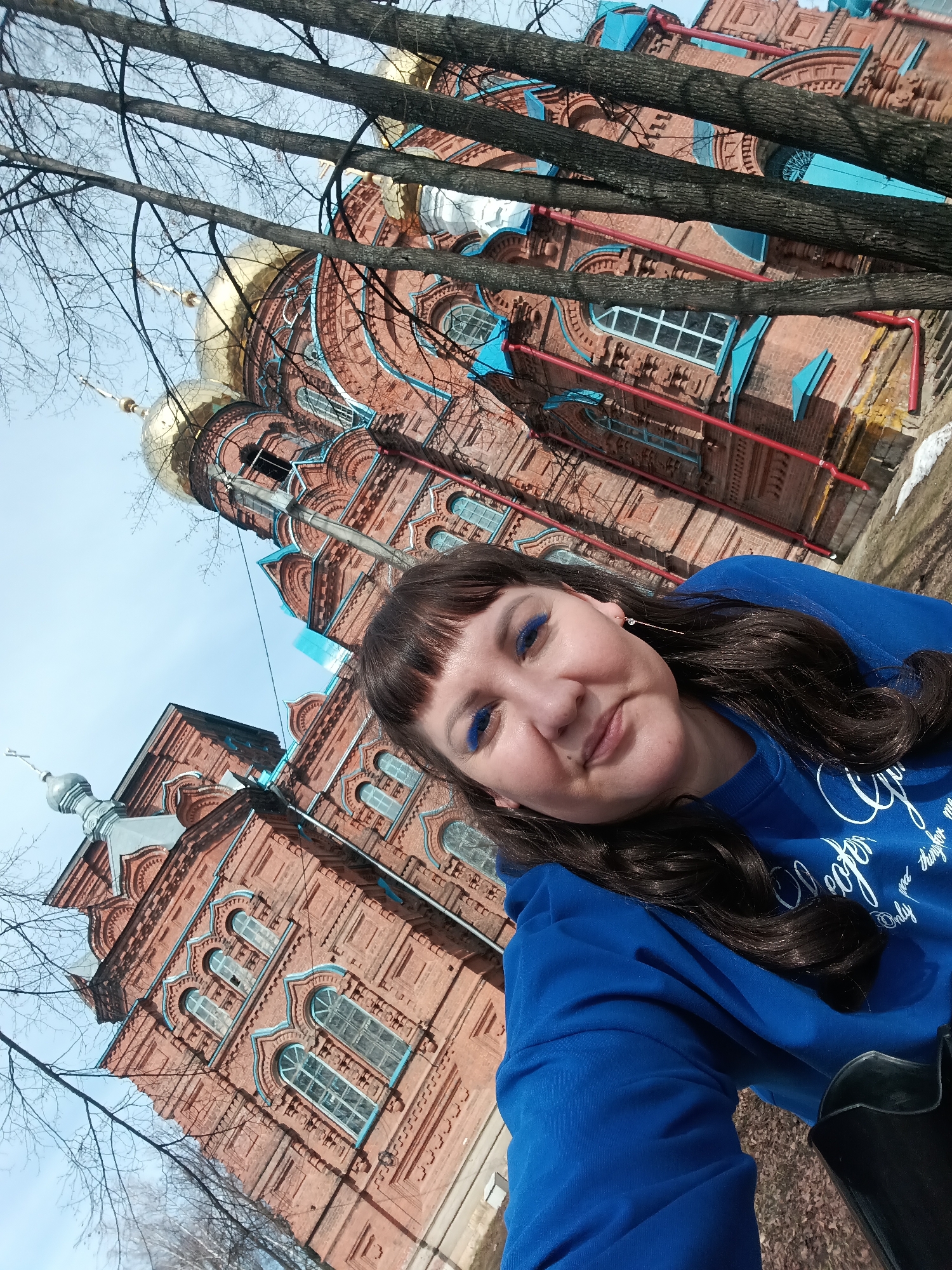 The image is a selfie of a woman with brown hair and blue eyeshadow in front of a large, ornate red brick building with gold and silver domes. Bare tree branches frame the top of the image. The building appears to be a church or cathedral, with multiple arched windows and decorative brickwork. The woman is wearing a blue sweatshirt. The photograph was taken in Osa, Russia.