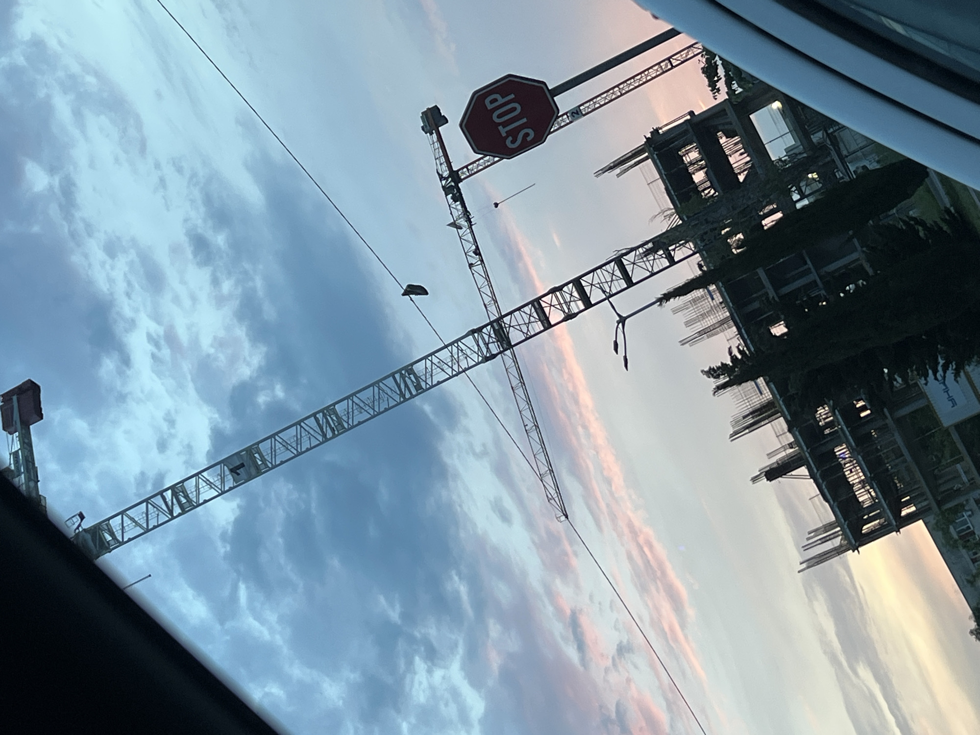 The vertical image, captured from inside a vehicle, depicts a construction site at either sunrise or sunset in Burgas, Bulgaria. The sky dominates the majority of the frame, transitioning from blue-grey clouds on the left to soft pink, orange, and light yellow hues on the right, indicative of the golden hour.
In the upper-middle section, two large tower cranes are prominently featured. A red octagonal stop sign, with the word "STOP" clearly legible upside down, is attached to a pole that appears to be part of one of the crane structures. The main crane's long jib stretches horizontally across the sky, with a small, dark, indistinct object hanging from one of its cables. Several thin, dark lines, likely power lines or cables, traverse the sky.
The top-left corner of the image shows a partial view of the vehicle's interior, including a dark window frame. Towards the bottom-right, partially constructed multi-story buildings with exposed concrete and rebar frameworks are visible, surrounded by dark tree foliage. Faint, unreadable text can be seen on a distant sign near these buildings. No individuals are discernible in the scene.