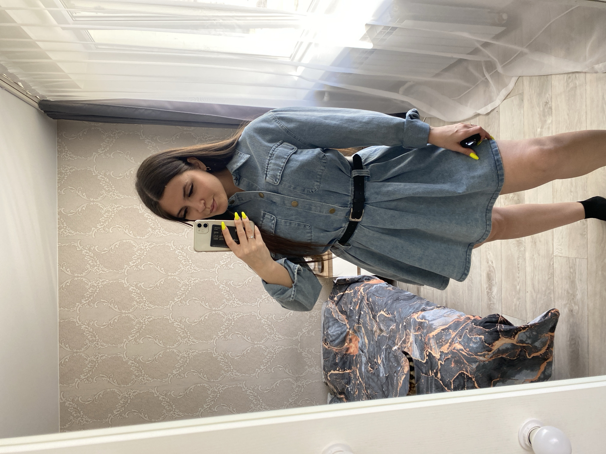 A woman is taking a mirror selfie indoors. The room has patterned wallpaper and wooden flooring. A chair with a marble-patterned cover is visible. Curtains hang by a window. The woman is wearing a denim shirt dress with a black belt and has yellow nail polish. The location is Chaikovsky, Russia.