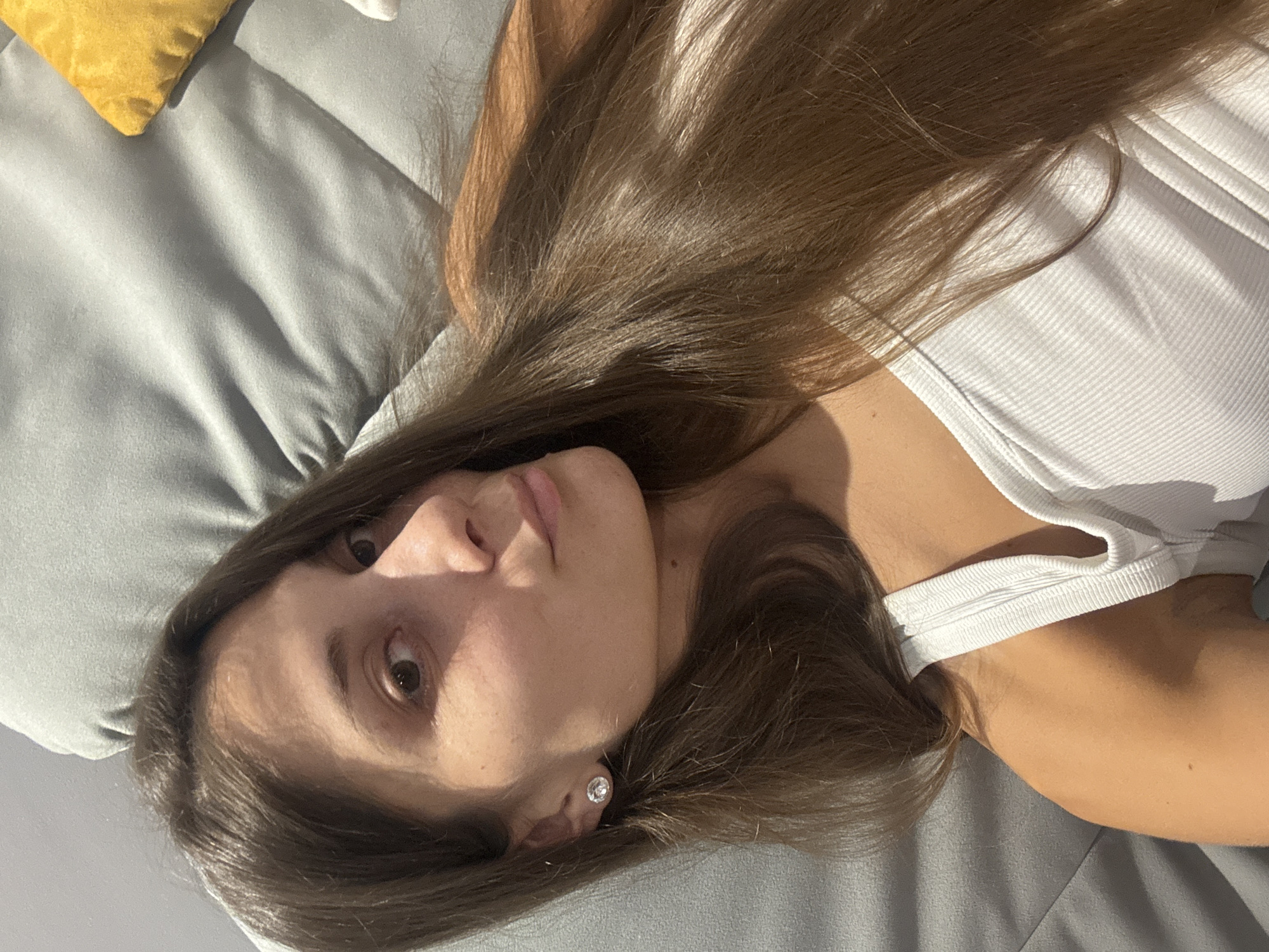 The image shows a young woman with long, brown hair lying on a gray surface, likely a couch or bed. She is wearing a white tank top and a stud earring in her left ear. Her head is turned towards the camera, and she is looking at it. Part of a yellow pillow is visible on the right side of the image. The lighting suggests it is indoors. There are no discernible landmarks that would place the image specifically in Burgas, Bulgaria.