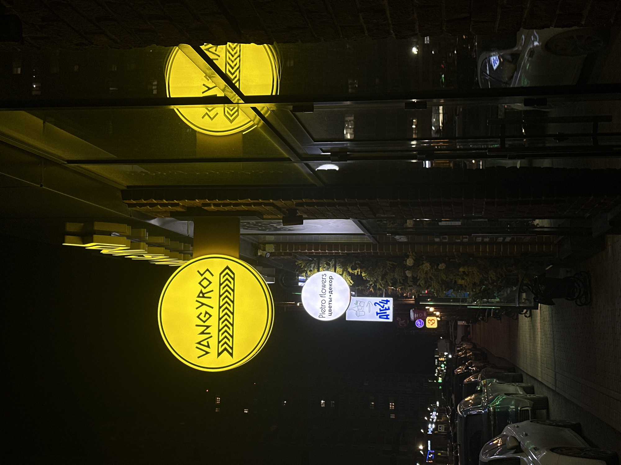 The image captures a nighttime street scene in Rostov-on-Don, Russia. Prominent signage indicates businesses along the street. A bright yellow, round sign displays "VANGYROS" vertically oriented. Nearby is a white, round sign for "Pietro flowers," with the text "цветы и декор" beneath. Adjacent to the floral sign is a rectangular sign "ATE4." Parked cars line the street, reflecting the ambient light. Distant building lights are visible in the background. The image is taken from a high vantage point, with the top portion showing a reflection of the "VANGYROS" sign.