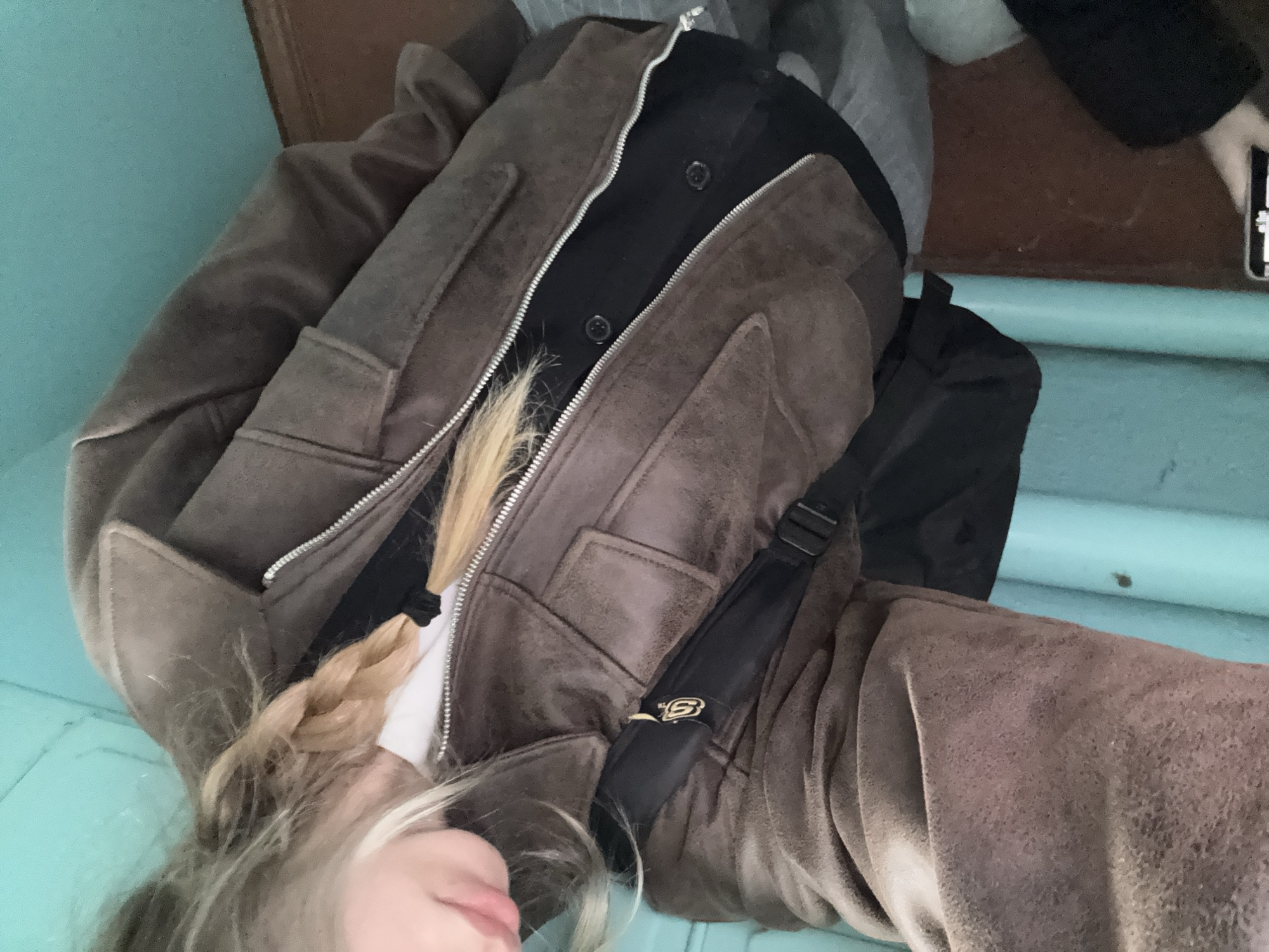 This media file features a close-up, rotated view of a person, likely a young individual, situated indoors. The main subject is an individual with long, light blonde hair, which is partially braided and falls over their left shoulder. Their face is partially visible, showing fair skin and a neutral expression.
The person is wearing a heavy, brown jacket with a textured, possibly suede or faux-suede, finish. The jacket has a prominent silver zipper that is partially unzipped, revealing a black collared shirt underneath with two visible black buttons. A white layer, likely an undershirt or t-shirt, is also visible beneath the black shirt. A black strap, possibly from a shoulder bag or backpack, crosses the person's torso. On this strap, a gold-colored emblem or logo, resembling a stylized 'S' or part of a brand name, is visible.
The setting appears to be an indoor space, possibly a stairwell or hallway, indicated by a light blue or turquoise-colored surface that occupies a significant portion of the lower and left side of the frame. In the upper right corner, a dark brown, possibly wooden, surface is visible, along with a hand holding a smartphone, suggesting another person or the subject themselves might be taking the photo. The overall lighting is consistent with an indoor environment. No specific time of day or weather conditions are discernible. The location context provided is Noviy, Russia, though there are no explicit visual cues within the image itself to confirm this.