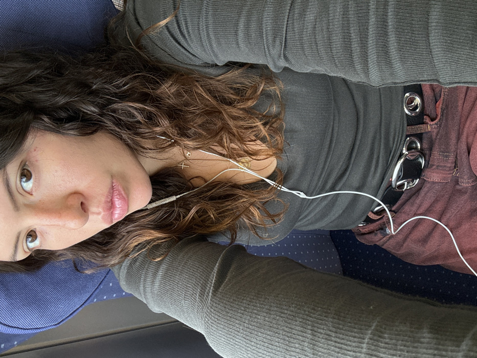 A female individual is depicted in a close-up, high-angle selfie, seated in what appears to be a public transport vehicle, likely a train or bus, indicated by the blue patterned seat behind and below her. She has long, wavy brown hair, brown eyes, and her lips are slightly pursed. She is wearing a dark gray or olive-green ribbed long-sleeved top and dark red or maroon-colored pants, secured with a black belt featuring a large silver-toned buckle and two silver eyelets. White wired earphones are in her ears, with the cord extending down her front. Multiple necklaces are visible, including one with a small silver-toned cross pendant and another with a round pendant. The individual is looking directly into the camera. The interior environment, consistent with public transport seating, offers no discernible visual markers specific to Reichertshausen, Germany, or any other precise geographic location.