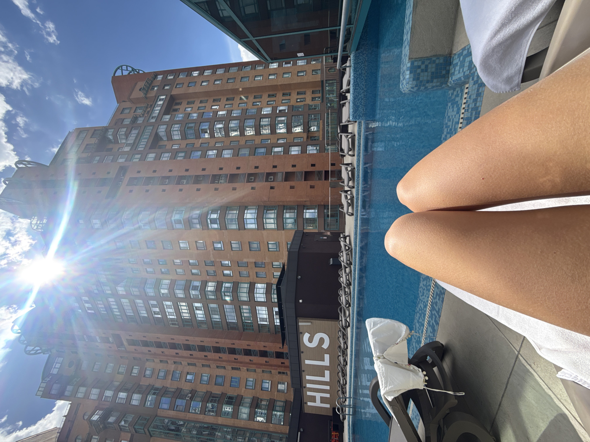 A person's legs are visible in the foreground, resting on a towel by a swimming pool. The pool water is a vibrant blue, with tiled edges. Behind the pool, a large, modern building with a brick facade and many windows dominates the background. The building's architecture is angular and imposing, with some sections appearing to jut out. The sky is a bright blue with scattered white clouds. The sun is shining intensely, casting a lens flare across the image and illuminating the building. On the side of one of the buildings, the word "HILLS" is visible in large white letters. The scene suggests a warm, sunny day, likely in an urban environment with access to outdoor leisure facilities. The perspective and composition create a sense of relaxation and enjoyment of the pleasant weather, potentially in Moscow, Russia, given the architectural style.