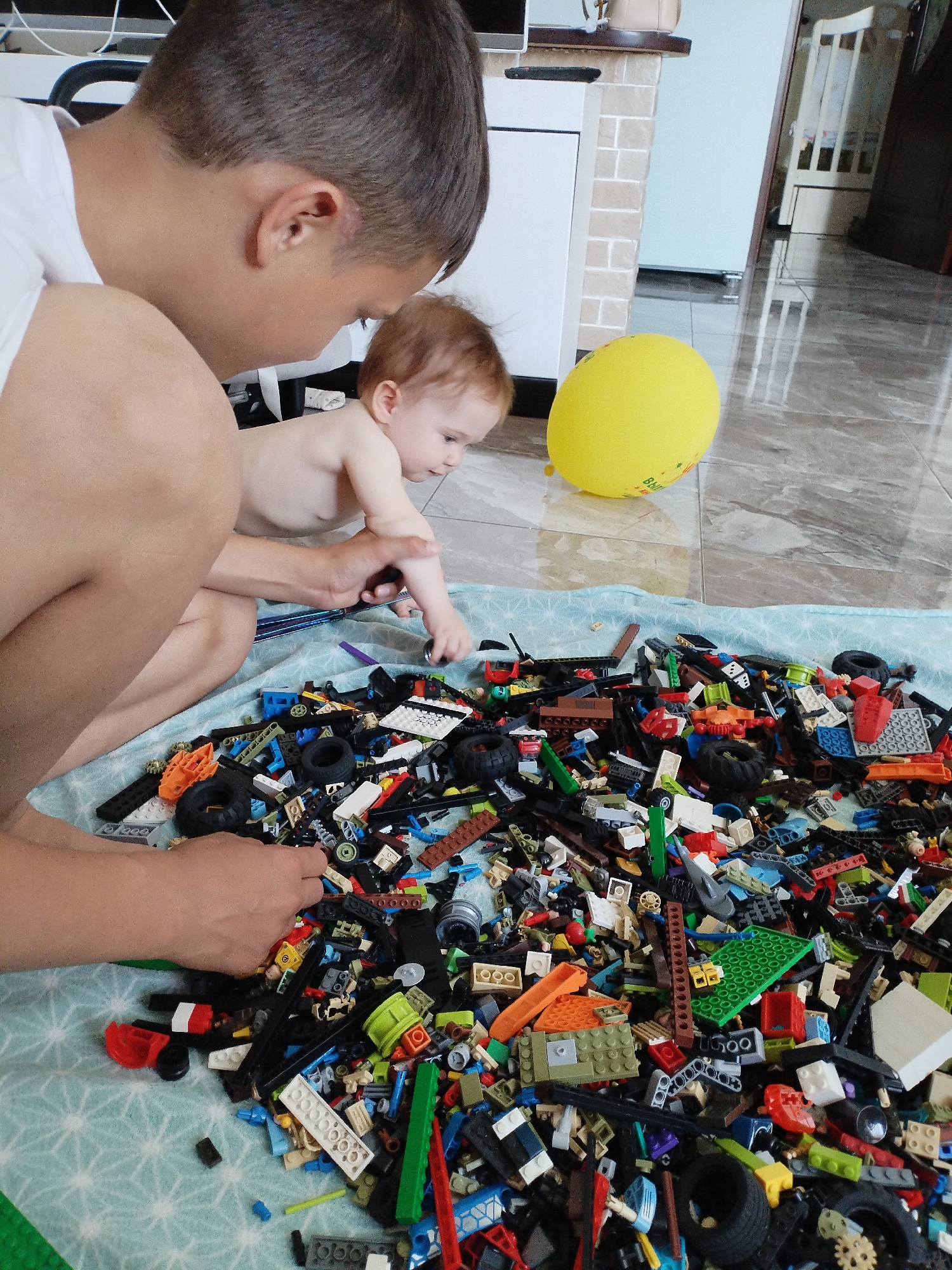 This image captures an indoor domestic scene in Vinogradny, Russia. In the foreground, an adult male, partially visible from the side and back, wearing a white t-shirt, kneels or squats on the left. His hands are engaged with a large pile of assorted building blocks, primarily Lego, scattered on a light blue mat with a subtle white pattern. To the adult's right, a baby, shirtless and with light brown hair, is on its knees, reaching into the pile of blocks with its right hand. The adult's left hand appears to be near the baby's left arm. The pile of blocks is substantial, containing a mix of colors, shapes, and specialized parts including wheels, plates, and bricks. Behind the baby, on a glossy light grey tiled floor, a partially deflated yellow balloon rests. The background reveals a room interior with white furniture, a textured wall section, and reflections on the floor showing elements such as a doorway and a white slatted chair.