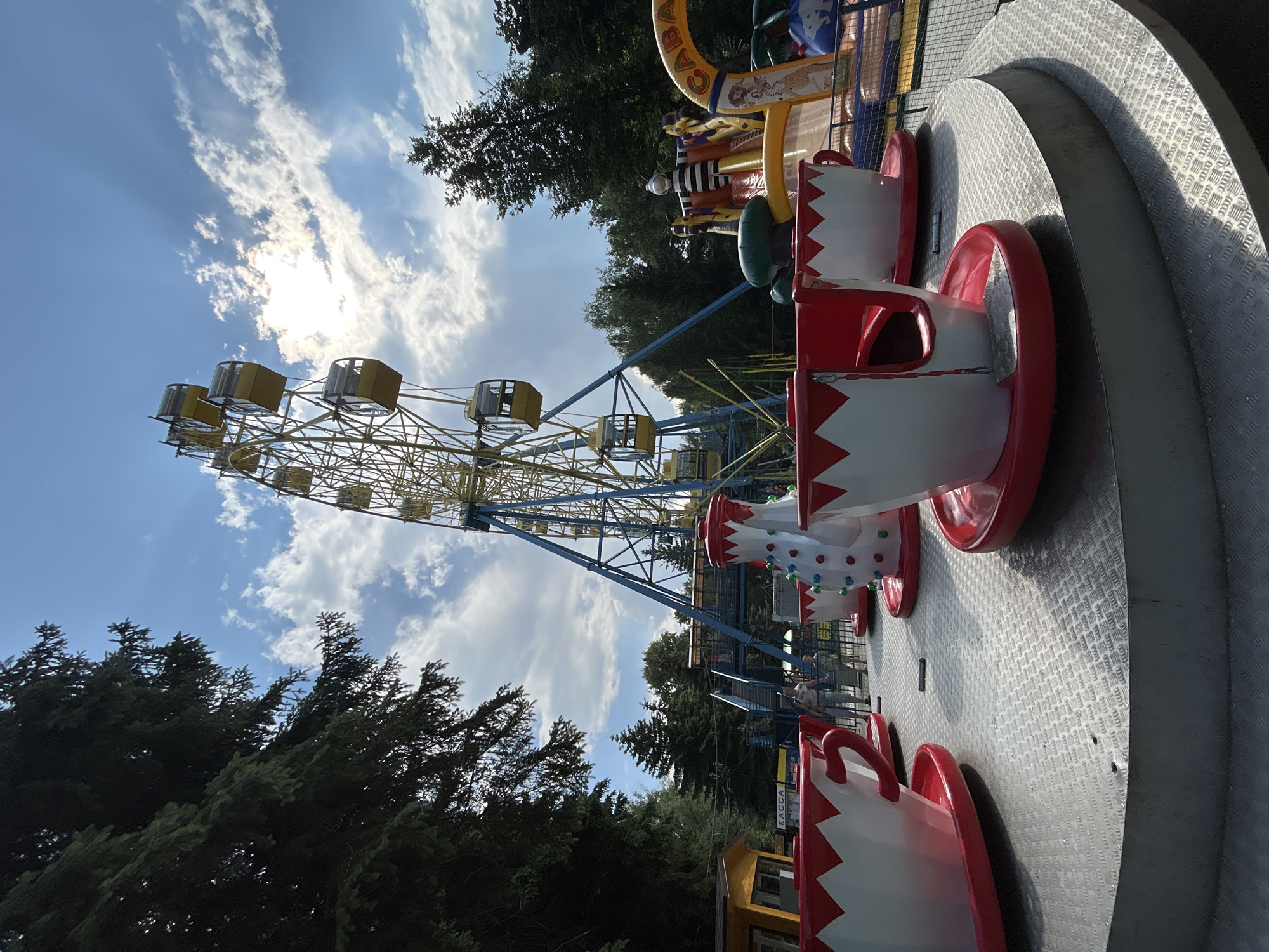 An amusement park ride featuring teacups is in the foreground. The teacups are white with red rims and saucers. Behind the teacups, a Ferris wheel with yellow gondolas is visible. The Ferris wheel is tall and stands against a blue sky with scattered clouds and sunbeams. Trees surround the amusement park. To the right of the Ferris wheel, there is a colorful inflatable slide with a character dressed in a black and white striped costume. In the middle ground, several people are walking near the ride. A small yellow building with a sign that says "КАССА" (KASSA, meaning "cashier" or "ticket office" in Russian) is located to the left of the teacup ride. The scene appears to be in an outdoor park setting in Yoshkar-Ola, Russia, judging by the Cyrillic signage and the architectural style of the Ferris wheel and ticket booth.