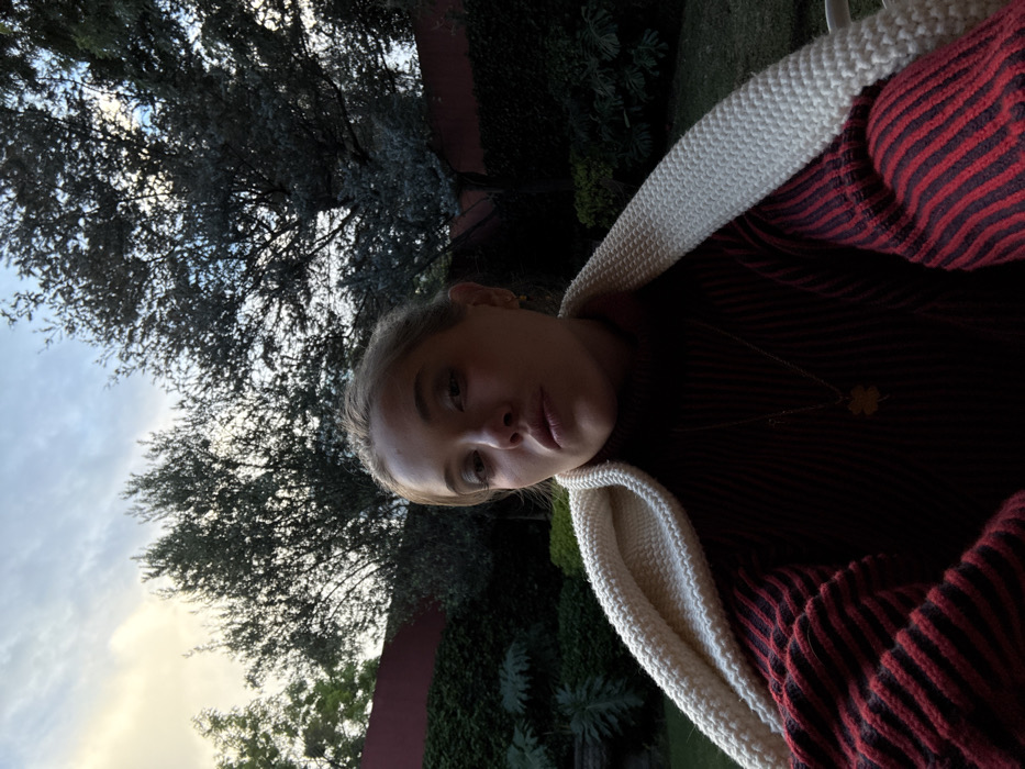 A person is centered in a tilted selfie with a tree, sky, and wall in the background. The person has fair skin, blond hair pulled back, and wears a red and dark blue striped sweater with a white crocheted shawl. They are also wearing a delicate gold necklace with a clover pendant. The background features a lush green tree against a cloudy sky on the left and a dark green wall and foliage on the right.