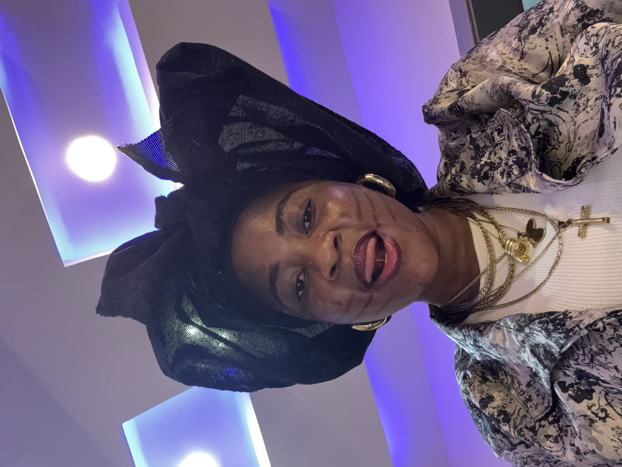 In Ajara, Nigeria, a low-angle medium shot captures a woman wearing a black headscarf and a patterned jacket over a white shirt. She has dark skin, prominent cheek markings, and is sticking her tongue out. Several gold necklaces with pendants adorn her neck. The background features recessed ceiling lights emitting a purple hue.