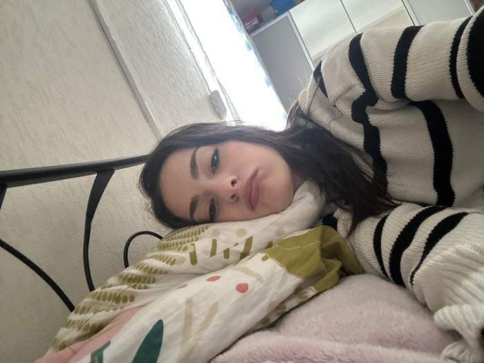 The scene is a residential room in Strasbourg, France, featuring a single female individual in a bed. The woman, with dark hair, lies on her left side, her head resting on a pillow adorned with an abstract pattern of geometric shapes in white, pink, green, and orange. She is partially covered by a multi-colored duvet with similar patterns and a pink, textured blanket. She wears a white sweater with thick black horizontal stripes. Her eyes are open, directed towards the camera, and her lips are slightly parted. A black metal bed frame is visible along the left edge of the image. The background consists of a light-colored, textured wall, and a white, reflective wardrobe or cabinet is partially visible in the upper right, illuminated by a bright light source, likely a window.