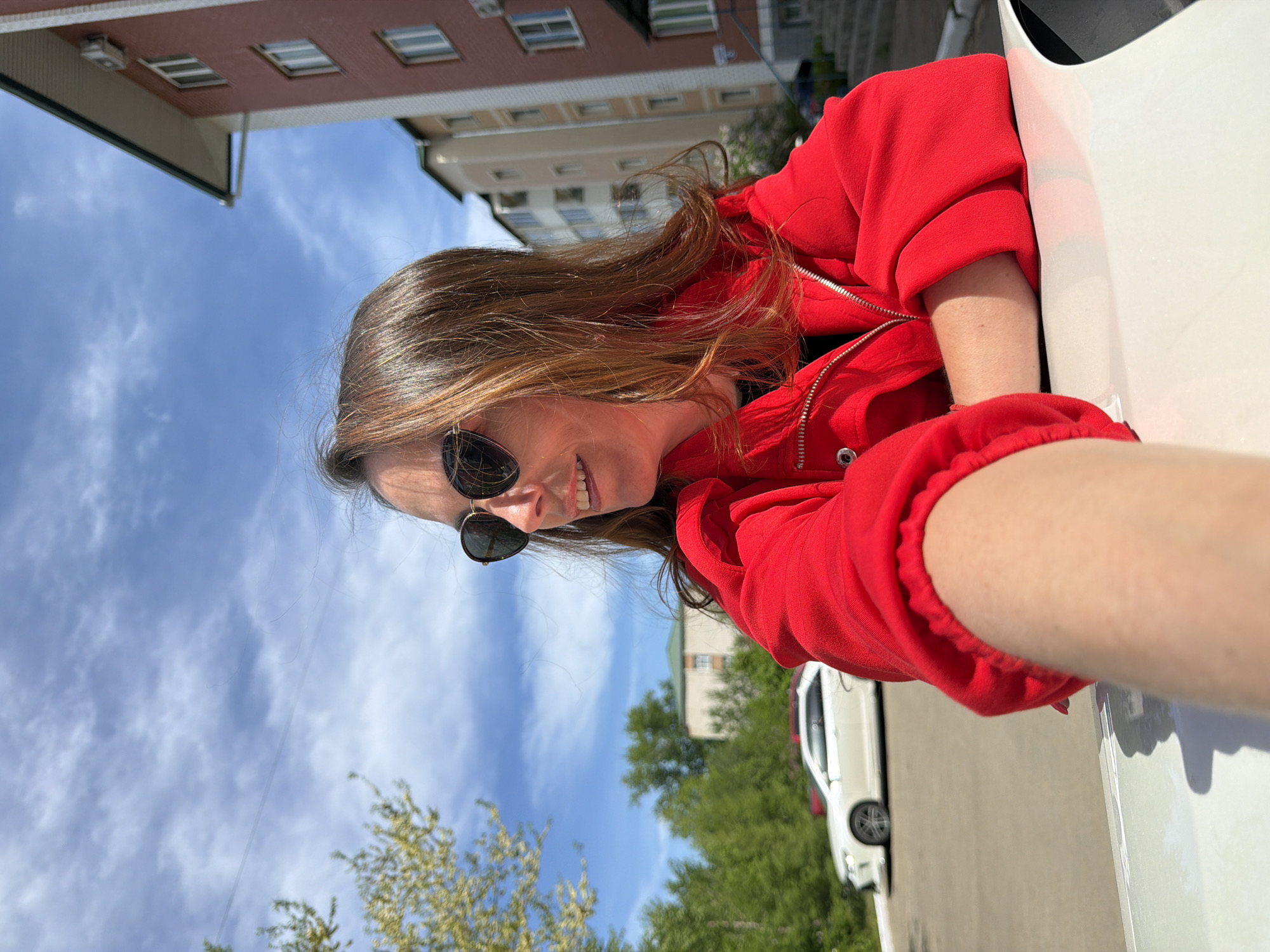 A woman with long, light brown hair, sunglasses, and a red hoodie is shown in a low-angle close-up shot from the waist up. She is leaning out of the driver's side window of a white car and smiling. The car appears to be parked on a street. In the background, there are buildings, trees, and a blue sky. The buildings suggest an urban environment, potentially in a city. It is impossible to confirm the image's location within the city of Birobidzhan, Russia.