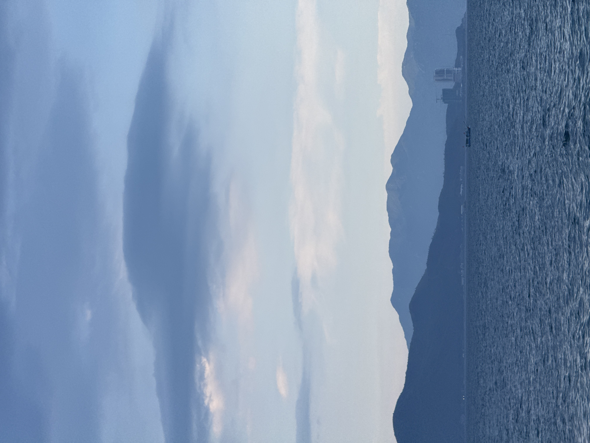 This is a landscape photograph taken from the sea, looking towards the coast of Nha Trang Ward, Vietnam. The image captures a serene coastal scene with several distinct layers.
In the foreground, the deep blue sea is visible, with gentle waves rippling across its surface. A small fishing boat is positioned in the middle ground, traversing the water. Beyond the sea, a series of layered mountains form the backdrop. The mountains are rendered in varying shades of blue and purple, indicating distance and atmospheric perspective.
On the coastline, nestled amongst the mountains, are signs of human development. A few buildings, including what appears to be a high-rise structure and construction cranes, are visible, suggesting ongoing development or urban presence. The sky above is a blend of soft blues and grays, with scattered clouds adding texture. One prominent, elongated cloud formation is centered in the upper portion of the image, adding a dramatic element to the sky. The overall impression is one of a vast, tranquil seascape meeting a mountainous, developing coastline under a diffused sky.