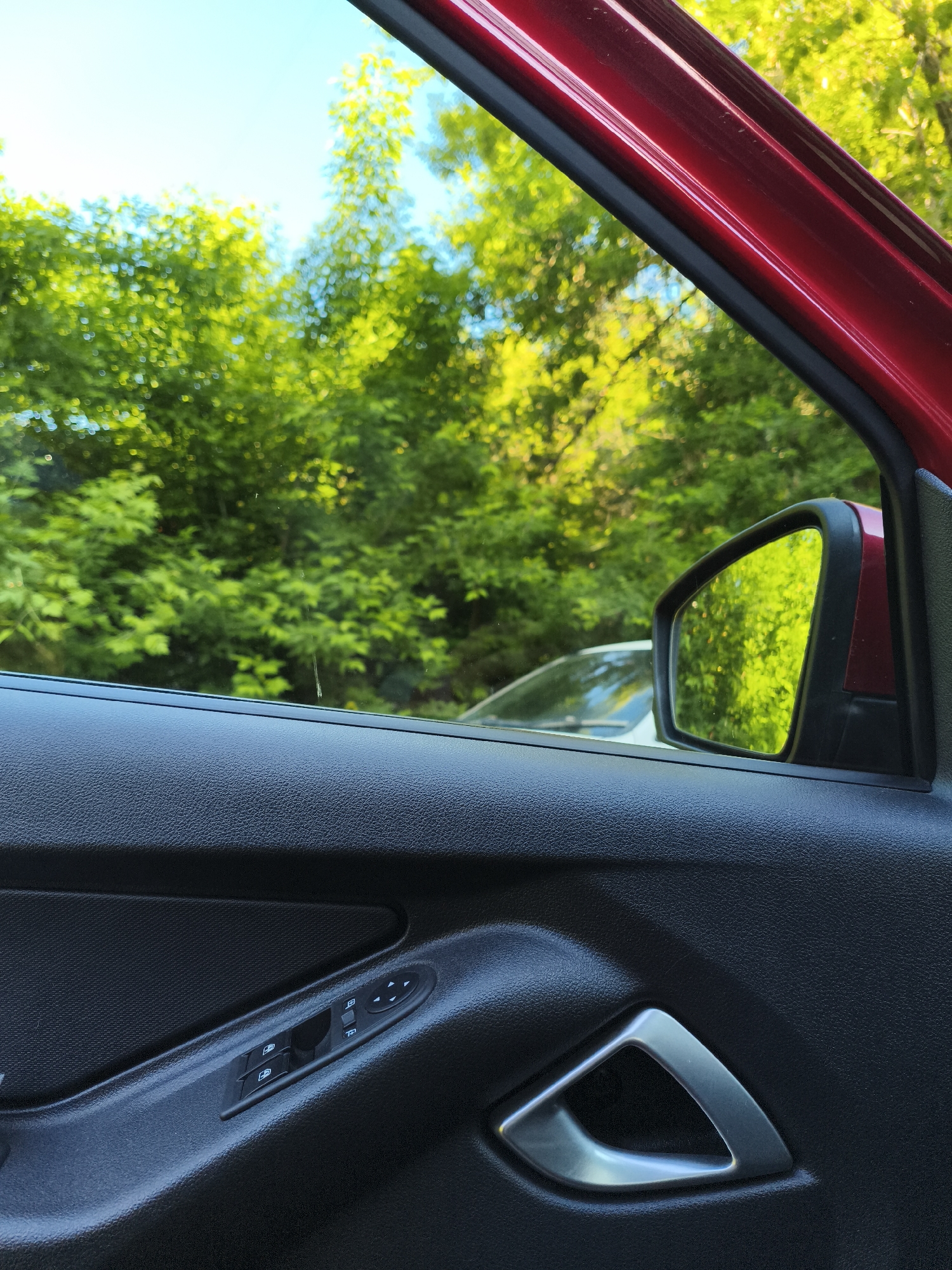 The media file captures a view from inside a red car, looking out through the open window on the passenger or driver's side. The scene is dominated by vibrant green foliage and a clear blue sky, suggesting a natural or park-like setting in Izhevsk, Russia.
In the foreground, the black, textured plastic interior panel of the car door is prominently featured. On this panel, a set of controls is visible: two buttons with icons for power window operation (likely front and rear) and a small joystick-like control panel for adjusting the side mirrors, marked with directional arrows. Below these controls, a sleek, triangular-shaped metallic silver door handle is embedded in the panel.
The top right corner of the image shows a sliver of the car's exterior, which is a shiny, deep red. The car's side mirror is also visible, reflecting the lush green trees from the background and partially showing the front of a white car positioned behind the red vehicle.
Beyond the car, the main subject of the exterior view is a dense canopy of trees with bright green leaves, heavily illuminated by direct sunlight. A clear, light blue sky is visible above the treetops. The bright light and clear sky indicate a sunny daytime setting. No people are visible, and no legible alphanumeric text is present, only graphical icons on the car controls. The image conveys a calm, bright outdoor moment from within a vehicle.