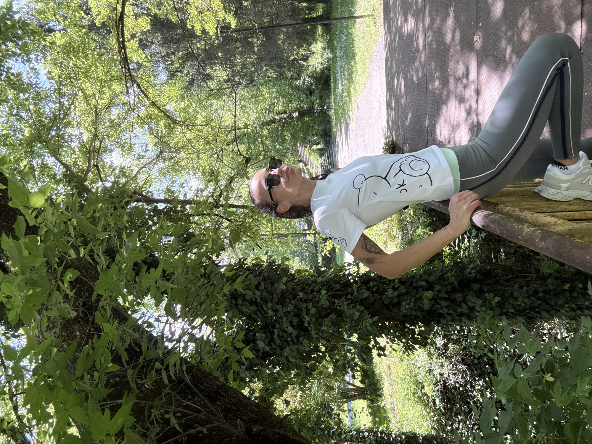 A woman with long, dark hair and sunglasses, wearing a white T-shirt with a cartoon graphic and olive green leggings with a white stripe, rests on a weathered, possibly wooden, bench in a park. The bench is surrounded by lush greenery, including trees and ivy-covered surfaces. A paved path, possibly part of a park walkway, is visible in the background. The scene appears to be in a park setting, judging by the trees, sunlight, and pathway. It may be from the city of Pleven, Bulgaria. The woman is holding onto the bench, suggesting she is either sitting or leaning on it. The image seems to capture a moment of relaxation in a natural environment.
