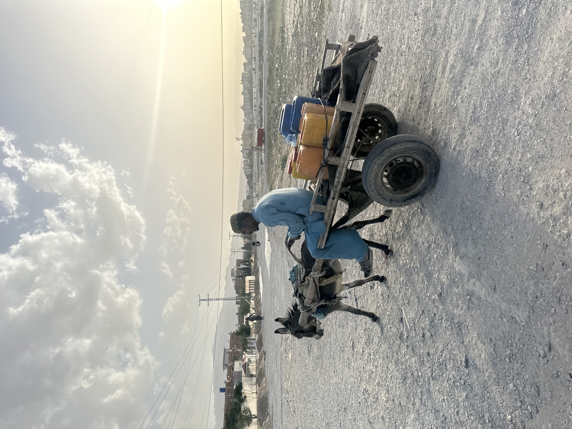 A man in a blue shirt and pants is riding a donkey, which is pulling a wooden cart with plastic containers. The cart has two wheels and is loaded with several blue and yellow plastic containers, possibly carrying water. The scene appears to be in Quetta City Tehsil, Pakistan, based on the surrounding landscape, architecture, and the presence of power lines. The ground is gravelly, and the background features buildings and mountains. The sky is bright with clouds.