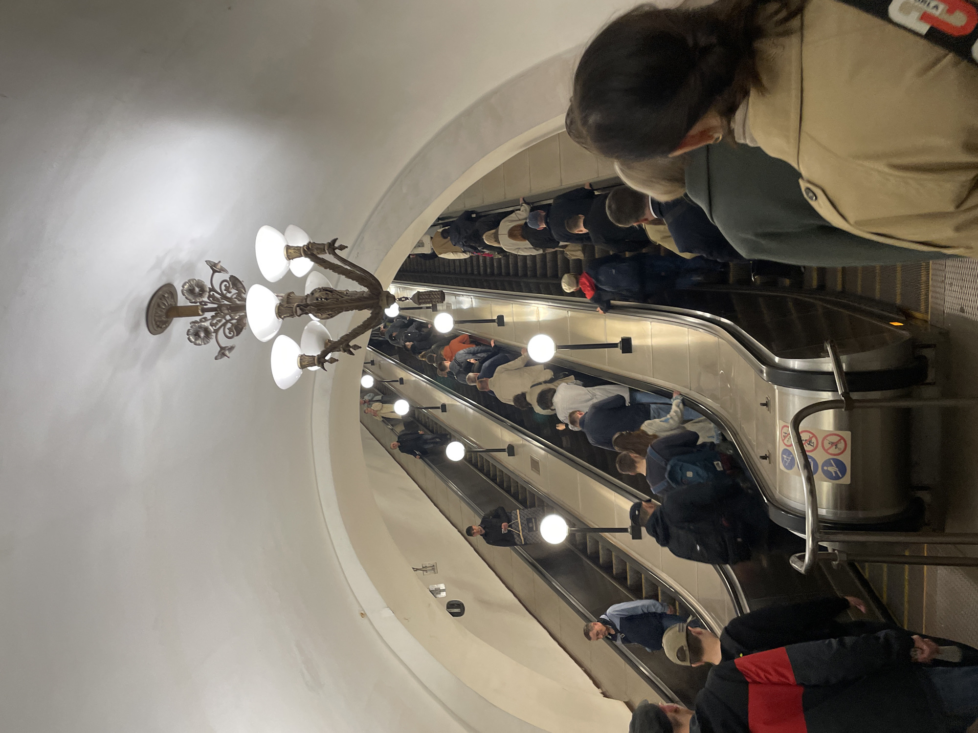 The image depicts a scene inside a Moscow metro station in Russia. Multiple escalators are visible, descending into the station. A chandelier hangs from the vaulted ceiling above the escalators. Numerous people are riding the escalators, most facing downwards. Some individuals are wearing casual clothing, while others are in more formal attire. Signs related to escalator safety are visible on the side. The environment is well-lit, and the architectural style suggests a grand, ornate design characteristic of Moscow's metro stations.