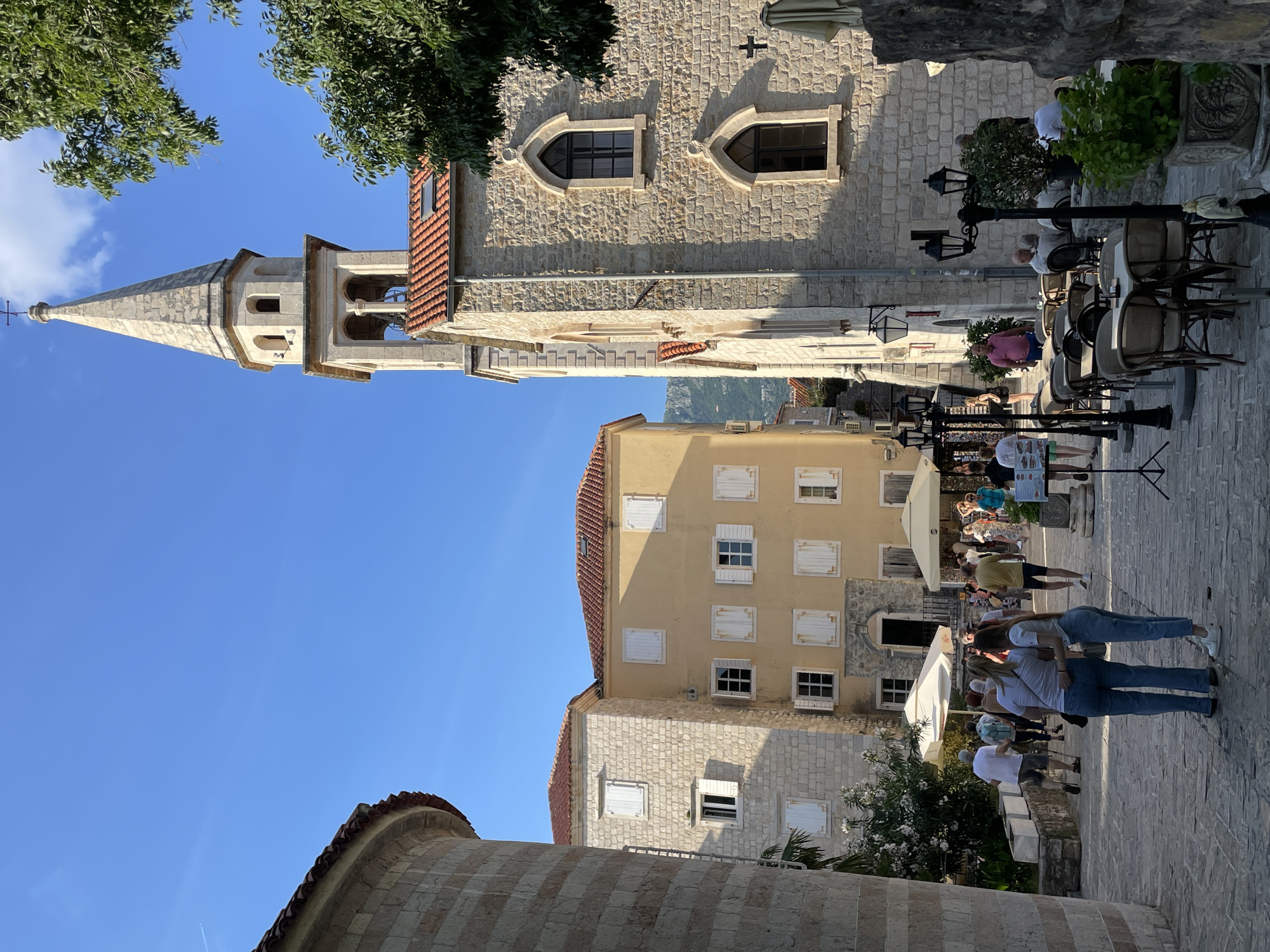 The image captures a scene in the city of Budva, Montenegro. The foreground features a rounded, striped building. Further back, a yellow building with white shutters rises behind a stone building. Tall, stone church with a pointed steeple dominates the left side of the frame. A sidewalk with people is visible, some appear to be tourists or visitors. Outdoor seating, including tables and chairs, is situated near a building, potentially a cafe or restaurant. Streetlights are interspersed among the seating area. The sky is bright and clear.