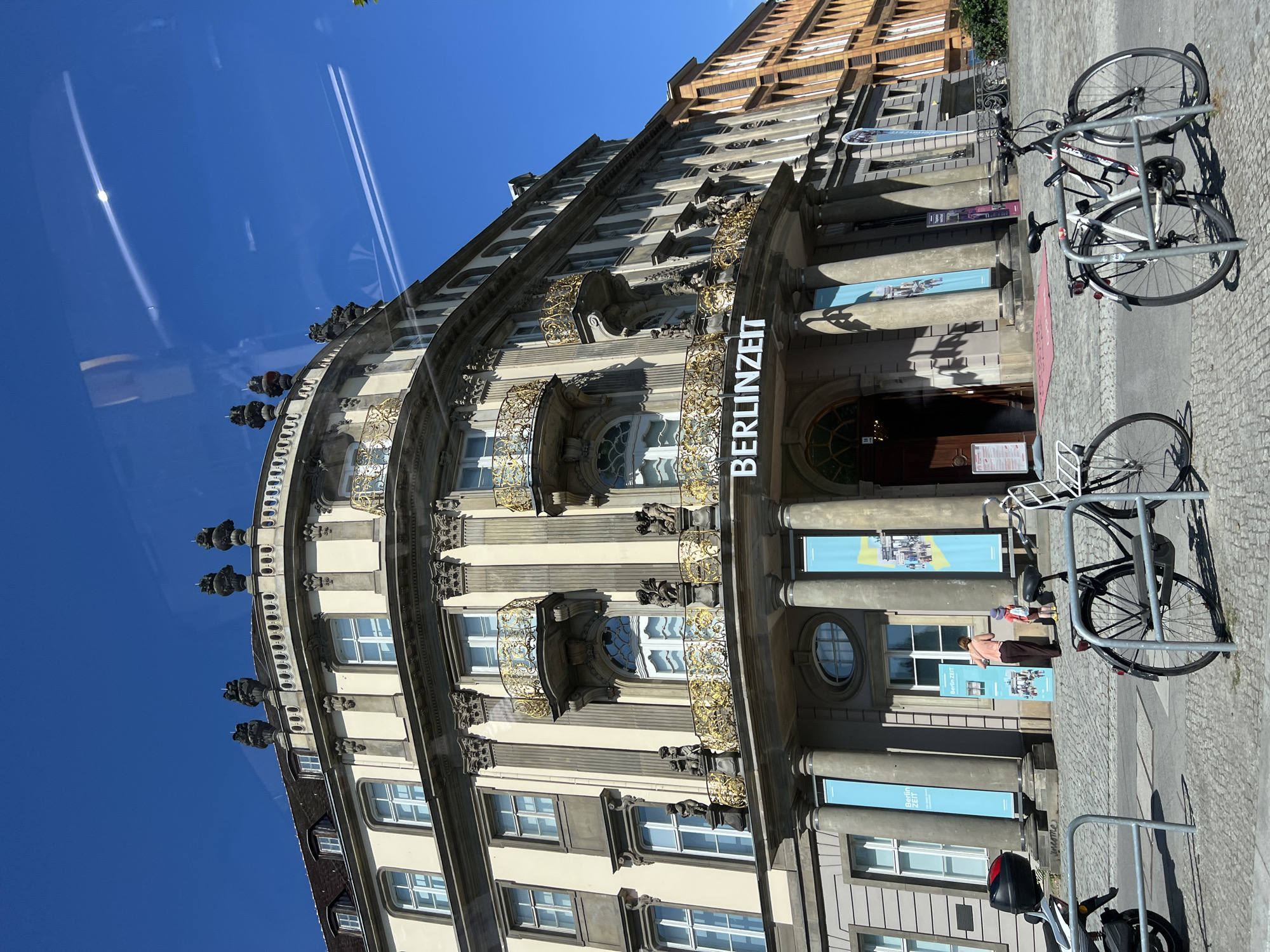 The image shows the facade of the "BerlinZeiT" museum, located in Berlin, Germany. The museum is housed in a grand, historic building with ornate architectural details, including classical columns, decorative balconies with golden railings, and statues. The building's exterior is a light cream color with dark trim around the windows and cornices. Two light blue banners hang vertically on the building's facade, one featuring text and the other an image, both appearing to be museum advertisements. In front of the museum, several bicycles are parked, and a red carpet leads up to the entrance. Two people, an adult and a child, are visible near the entrance, standing by one of the banners. The sky is clear and bright blue, with some faint contrails visible from an airplane. To the right of the main building, another building with an orange facade is partially visible. The ground in front of the museum is paved with cobblestones.