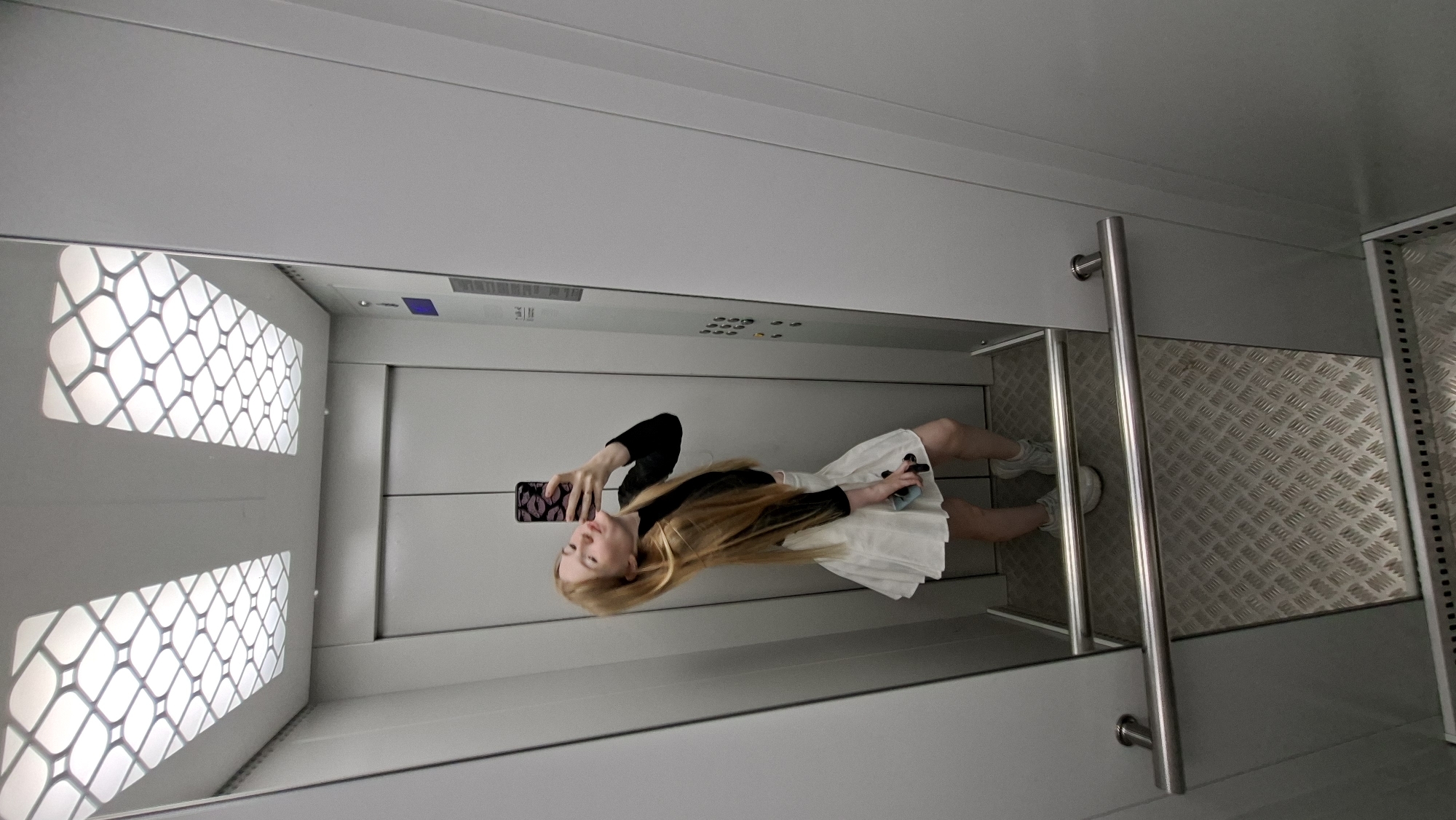 A young woman with long blonde hair is taking a selfie in an elevator. She is wearing a black long-sleeved shirt and a white pleated skirt. She is holding a phone with a kiss-patterned case and sticking out her tongue. The elevator has grey walls, a patterned ceiling light, and a metal floor. A mirror reflects the scene. There is an elevator control panel on the right wall. The scene is set in an elevator, likely in a public building, but the specific location in Chaikovsky, Russia, cannot be determined from the visual information.