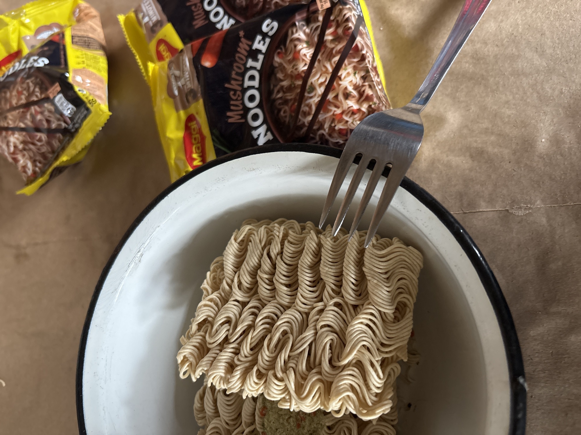 The image captures a close-up, overhead view of instant noodles being prepared. The primary subjects are two yellow and black packets of "Maggi Mushroom Noodles" visible in the background, with one packet partially opened. In the foreground, a white enamel bowl with a black rim holds a block of uncooked, wavy instant noodles. A silver-colored metal fork rests on top of the noodles, with its tines partially inserted.
Underneath the top layer of noodles in the bowl, a greenish sachet of seasoning powder is discernible. The setting appears to be an indoor surface, possibly a light brown paper bag or a rustic table, which serves as the background for the items. The lighting is bright, suggesting it is daytime.
Visible text on the noodle packets includes "Maggi", "Mushroom Noodles", and the word "NOODLES" prominently displayed. Another part of a label shows "5 MINUTES". The overall scene depicts the initial stages of preparing a meal of instant noodles.