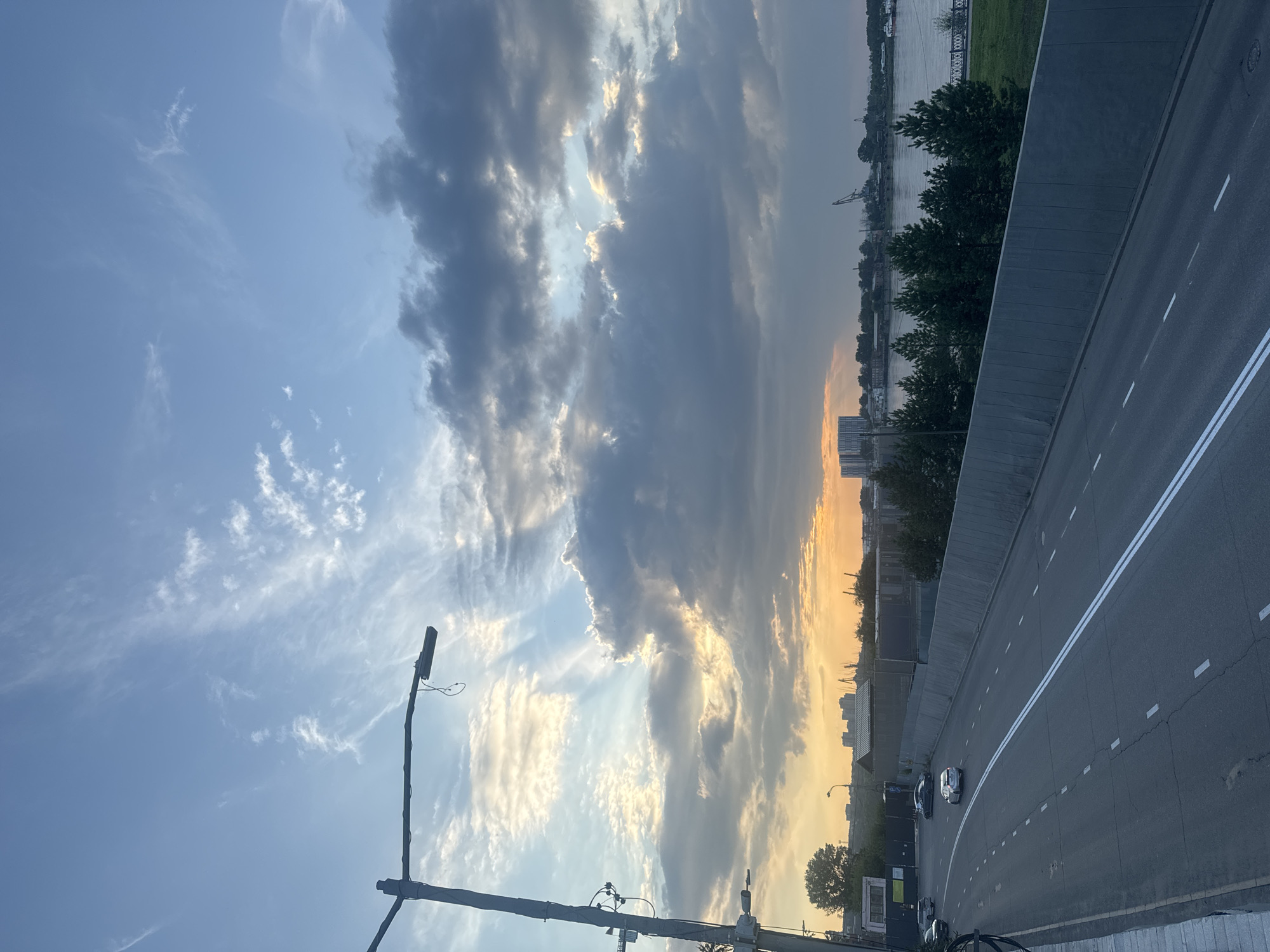 The image depicts a road leading into a city at sunset. The sky is filled with dramatic clouds, some dark and heavy, others illuminated by the setting sun, creating rays of light. A streetlamp stands prominently in the foreground on the left. The road itself is paved and marked with white lines, with a few cars visible. To the right of the road, there's a concrete wall and then a line of evergreen trees. Beyond the trees, a body of water, likely a river, is visible, with buildings and industrial cranes on the far bank. The city skyline in the distance is silhouetted against the warm hues of the sunset. The overall impression is an urban landscape transitioning into evening. While the image itself does not provide explicit markers of Irkutsk, Russia, the description of the scene is consistent with an urban setting along a river, which is characteristic of Irkutsk.
