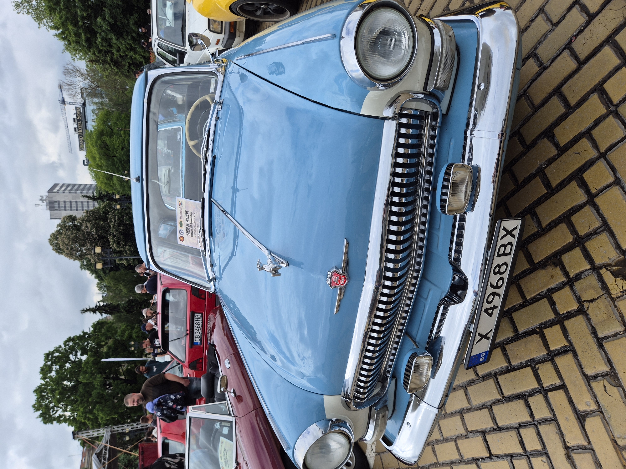 The image shows a vintage light blue car parked on a paved surface, with a focus on the front of the vehicle. The car has a prominent chrome grille and bumper, a hood ornament, and a license plate that reads "X 4968 BX" with "BG" (Bulgaria) underneath. A sign is visible on the car's windshield. A red car is partially visible to the left of the blue car, and people are standing nearby, suggesting a car show or gathering. In the background, one can see buildings and trees, including signs for "GRAND HOTEL SOFIA" and "CLASSIKS." This indicates that the location is most likely in the city of Sofia, Bulgaria, near the Grand Hotel Sofia.