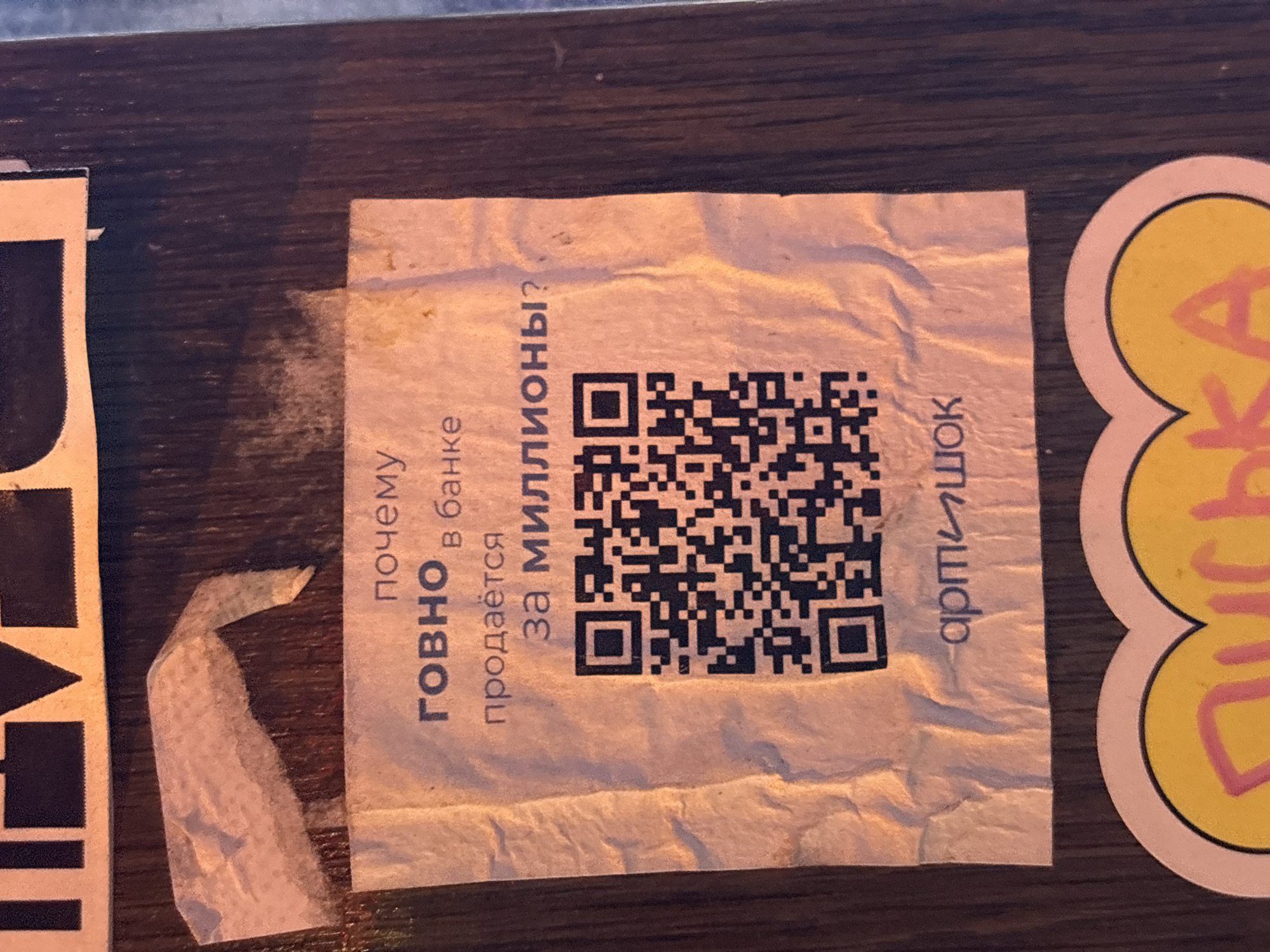 A rectangular, off-white sticker, appearing somewhat wrinkled and worn, is affixed to a dark, wooden-textured surface. The sticker prominently features a QR code in its center.
To the left of the QR code, vertically oriented Russian text reads: "почему ГОВНО В банке продаётся за МИЛЛИОНЫ?" This translates to "Why is SHIT sold in a bank for MILLIONS?", with the word "ГОВНО" (SHIT) appearing in a larger, bolder font. To the right of the QR code, also oriented vertically, is the Russian word "артИШОК" (artICHOKE), likely a brand or creator name.
The underlying surface is dark brown with a visible wood grain, suggesting it could be a table, counter, or wall. To the left of the main sticker, a partial black-and-white sticker is visible. On the right, a segment of a circular sticker with a bright yellow center and a pink border is present, showing partial pink text. The scene is static, devoid of people or activity, and appears to be an indoor setting, possibly in Saint Petersburg, Russia, where such stickers might be found on public or commercial furniture. The lighting is ambient, highlighting the textures of the materials.