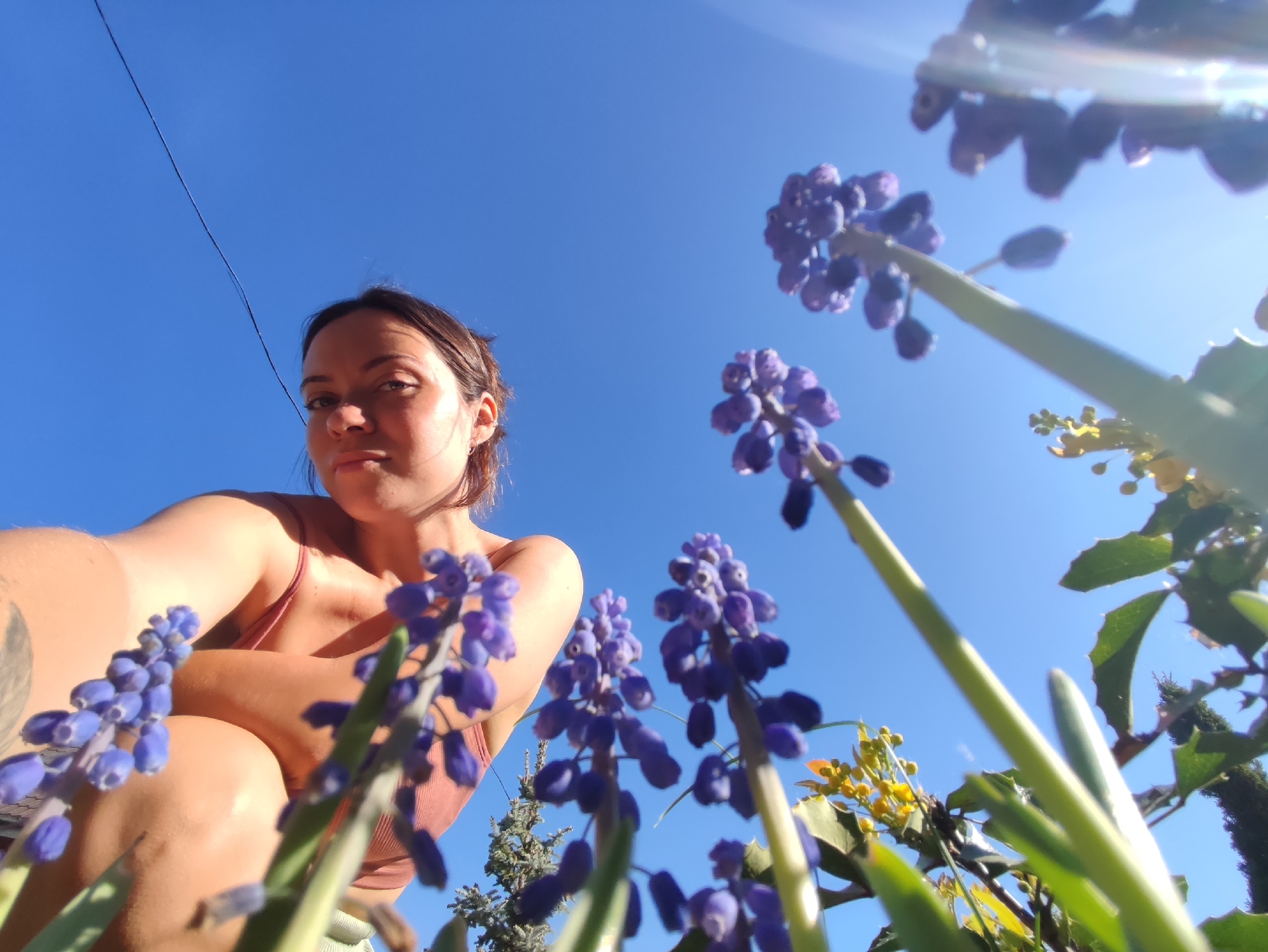 A woman is positioned to the left, looking at the camera. She has brown hair pulled back and is wearing a pink tank top. To the right and in front of her, there are several clusters of blue flowers, potentially grape hyacinths, angled upwards towards the bright blue sky. Some yellow flowers are visible behind the blue ones. A thin cable cuts across the upper left corner of the sky. The location is presumed to be Фоки, Russia.