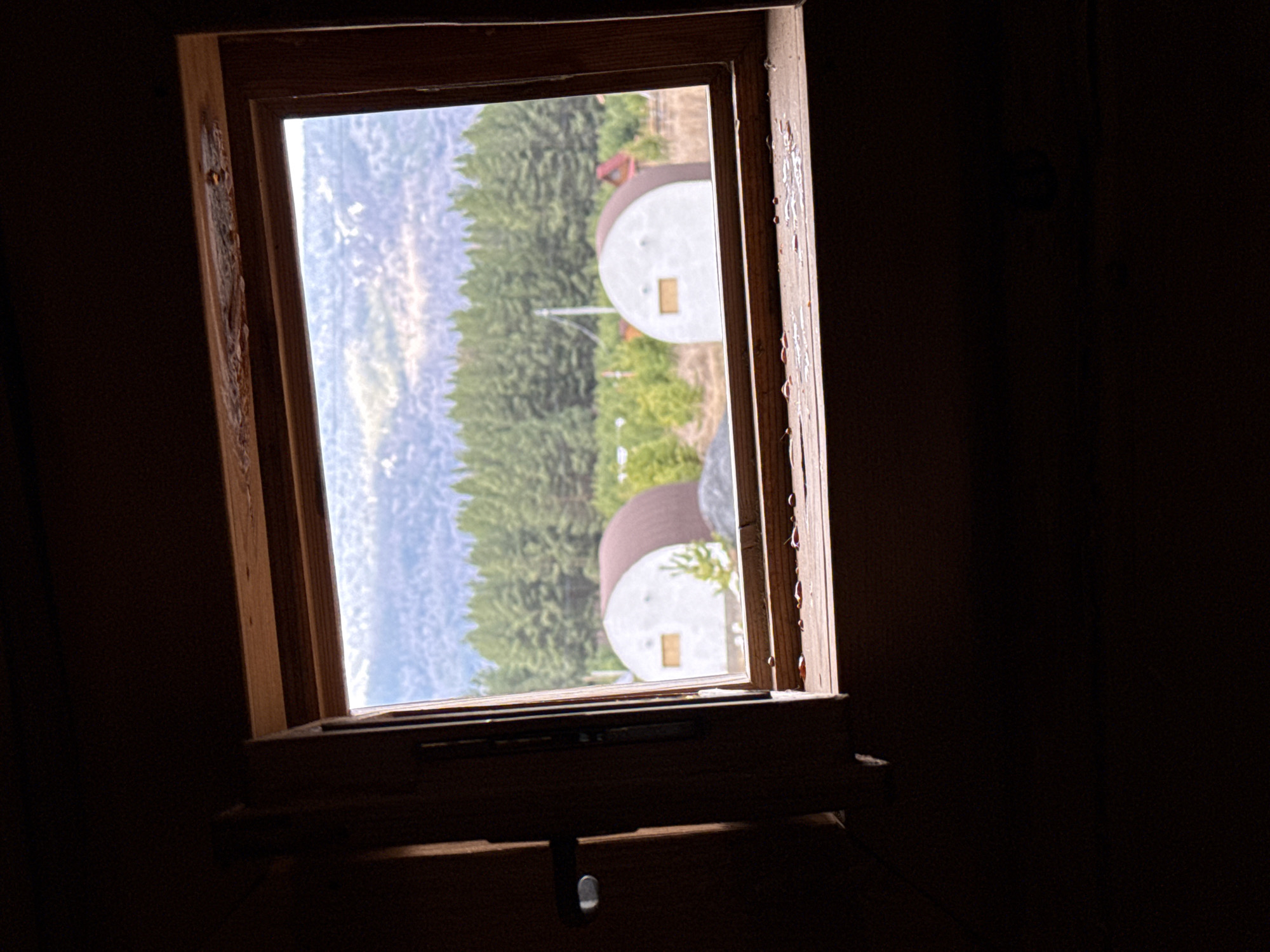 The image depicts a view through a wooden-framed window, likely from inside a dark room. Outside the window, the landscape includes a hillside with coniferous trees and two white, domed structures with brown tops, reminiscent of yurts. The scene is rotated, causing the sky to appear on the left and the structures to be oriented sideways. The window frame shows signs of wear, with drips or resin visible on the right side. Below the window is a sill with what appears to be a metal latch. The city name and country were added with editing.