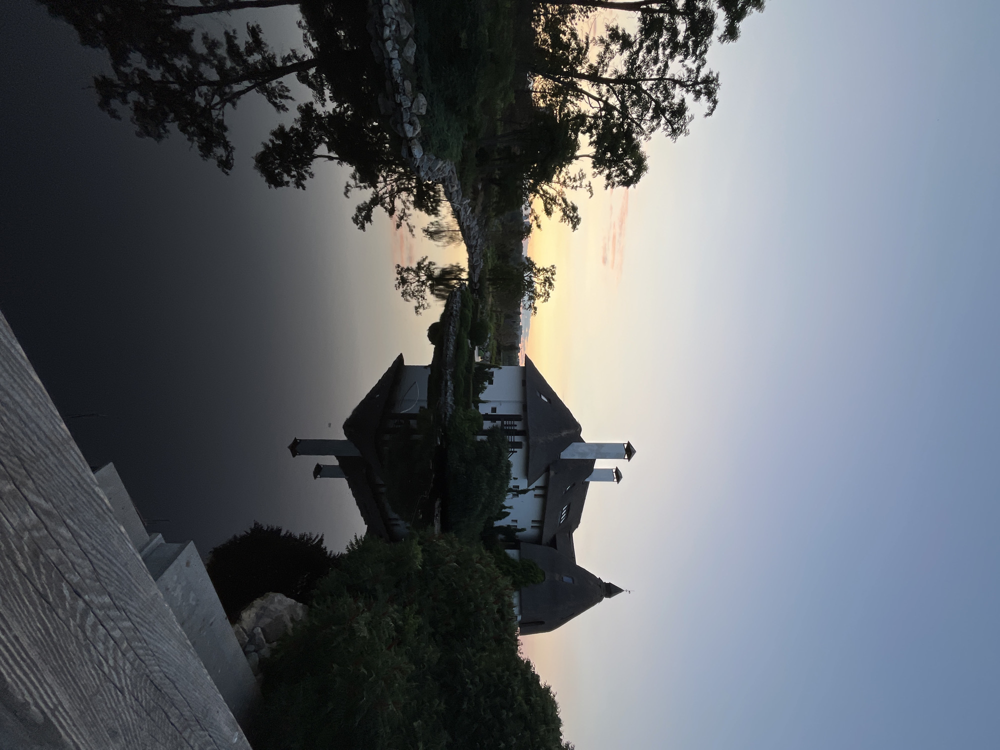 The image depicts a serene twilight scene by a tranquil body of water, likely a canal or small river, in the city of Dnipro, Ukraine. A large, multi-story building with a distinctive thatched roof and prominent chimneys stands on the right bank, partially obscured by lush green foliage. The building's white facade contrasts with the darker surroundings. The calm water perfectly reflects the building and the silhouettes of trees lining the left bank, creating a symmetrical and peaceful composition. The sky above transitions from a soft pale blue at the top to warmer hues of pink and orange near the horizon, indicating the end of sunset. A wooden railing or structure is visible in the foreground on the lower right, suggesting a viewing platform or walkway. No people are present in the frame. The overall atmosphere is one of quiet natural beauty enhanced by the unique architecture.