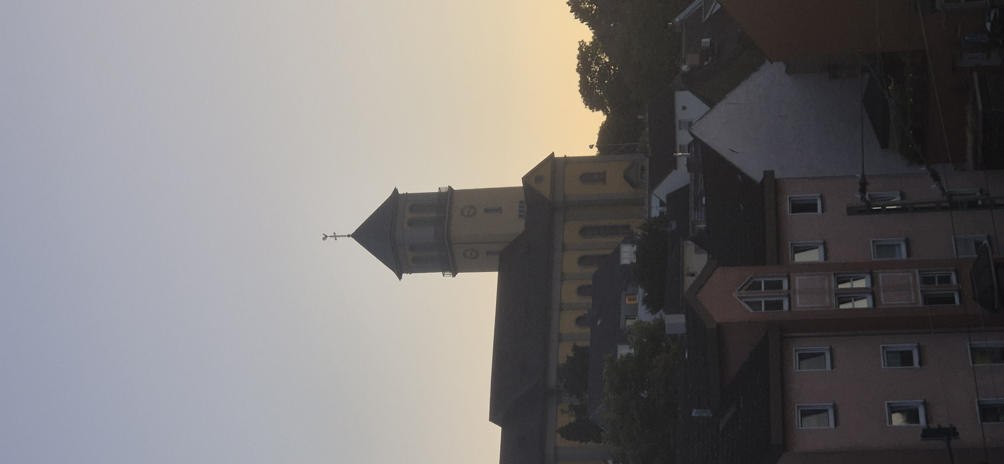 This is a vertical, medium-shot photograph of a yellow church with a dark roof and a tall, square bell tower topped with a steeple and a weather vane. The church is located in the city of Mainz, Germany. The church is situated on a hill and is surrounded by several residential buildings of varying heights and colors, including pink, white, and dark-colored structures. Trees are visible behind and around the buildings, and the sky above is a pale, gradient yellow and blue, indicating either sunrise or sunset. The scene is devoid of people and focuses on the architecture and the overall landscape.