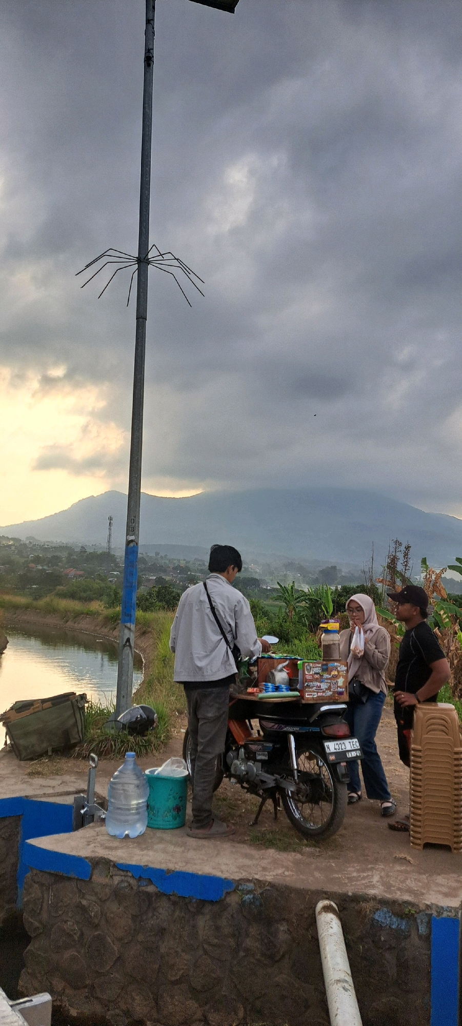 In Sumber Gedang, Indonesia, three people are gathered around a motorcycle with a mobile food cart attached. The scene appears to be outdoors with a body of water to the left and a mountainous landscape in the background under a cloudy sky.
The person on the left, facing away from the camera, is wearing a light-colored jacket and dark pants. They are interacting with the food cart. In the center, a person wearing a hijab and a light brown jacket is holding a snack and appears to be eating. To the right, a person in a black shirt and black baseball cap stands with their hands on their hips, observing the scene.
The food cart is mounted on an orange motorcycle, displaying various food items and containers. In the foreground, there are two large water jugs and a green bucket. Behind the people and the motorcycle, a stack of brown plastic chairs is visible. A large bag or backpack is placed on the ground to the left of the water jugs. A tall pole with a spider-like metal structure at the top stands to the left of the group, adjacent to a canal. The area is characterized by grassy banks and some vegetation. The overall atmosphere suggests a casual outdoor gathering or business transaction.