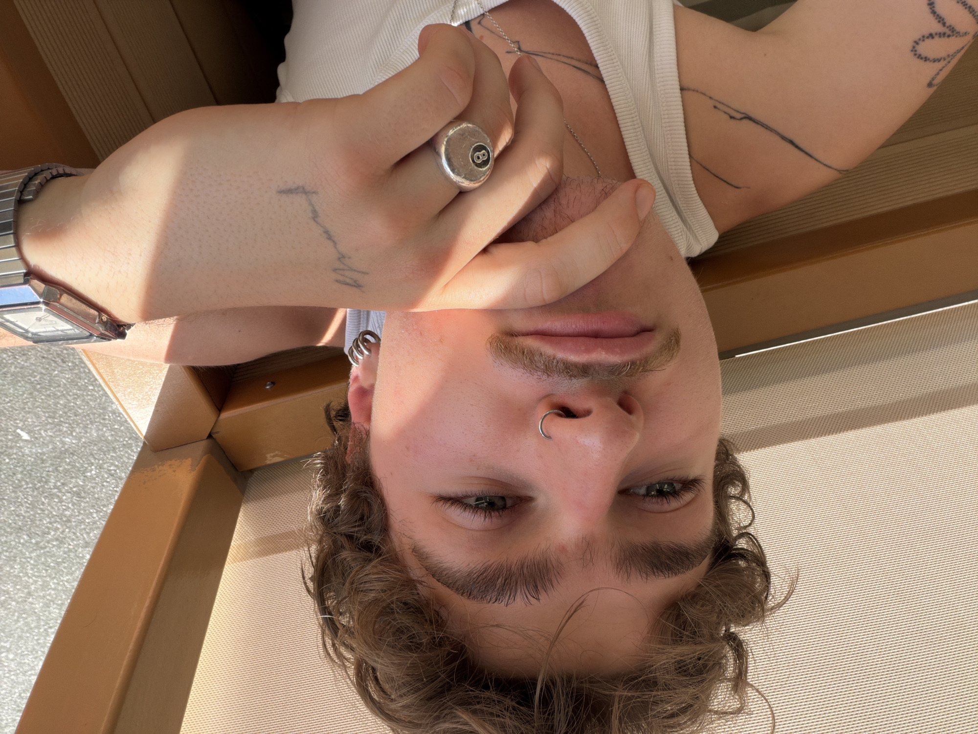 A man with curly brown hair and a nose piercing is visible in an upside-down, close-up photograph. He has light skin, green eyes, and a mustache. He is wearing a white tank top and a silver chain necklace. His left hand is raised, displaying a silver ring with an "8" on it, and a faded tattoo on his wrist. A silver watch is visible on his left wrist. Another tattoo is seen on his left arm. The man is sitting on a tan chair or bench with striped fabric. There is no indication of the location being Belgrade, Serbia.