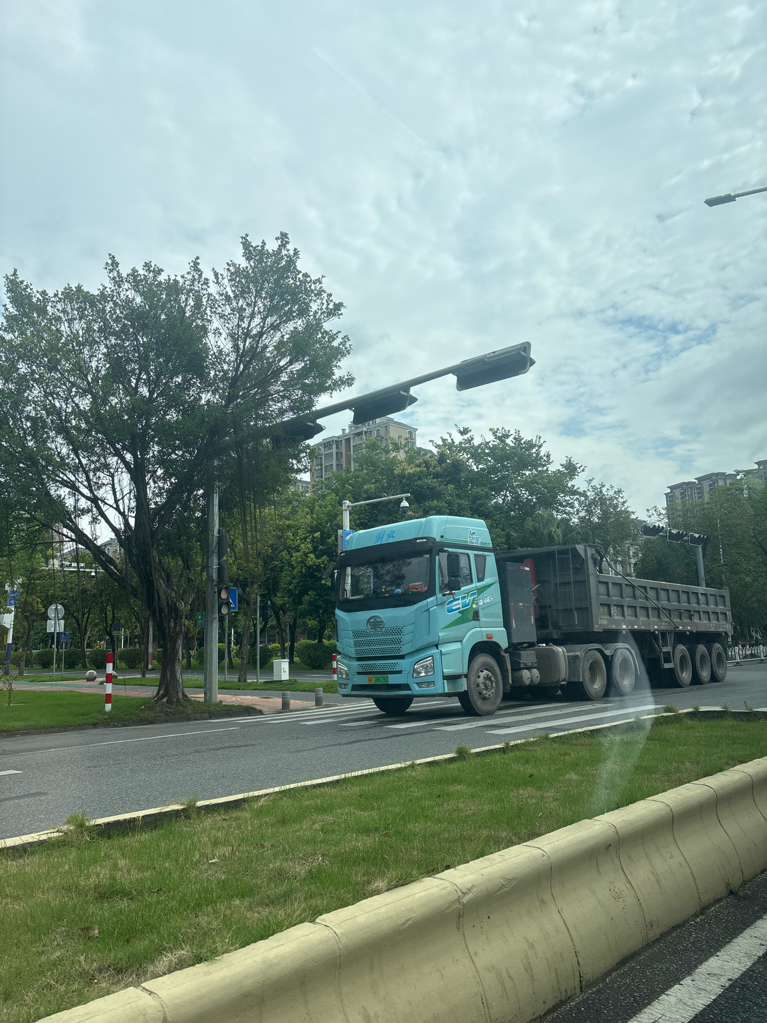 A light blue dump truck with a dark gray trailer is traveling on a road in Sanshui District, China. The truck has the license plate "R 096280". The truck is in the right lane of a multi-lane road, and there is a crosswalk in front of it. To the left of the road, there is a grassy area with a large tree and several traffic lights. In the background, there are apartment buildings and more trees. The sky is cloudy.